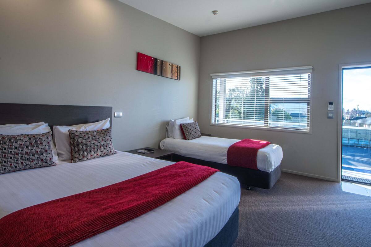 Une chambre d’hôtel avec deux lits, une fenêtre avec des stores et une œuvre d’art rouge accrochée au mur.