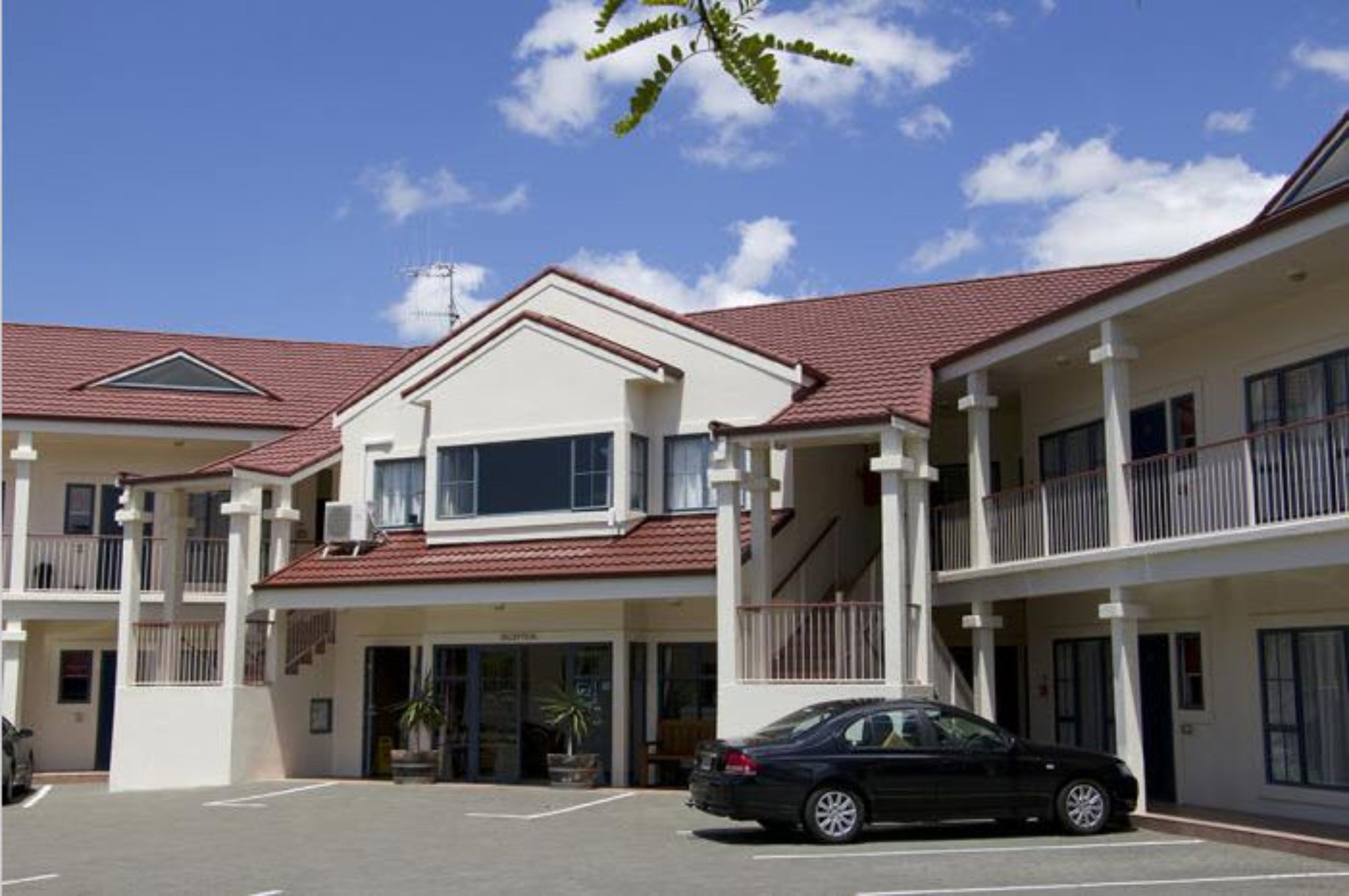 Um edifício de dois andares com telhado de telhas vermelhas, sacadas e um carro estacionado na frente.