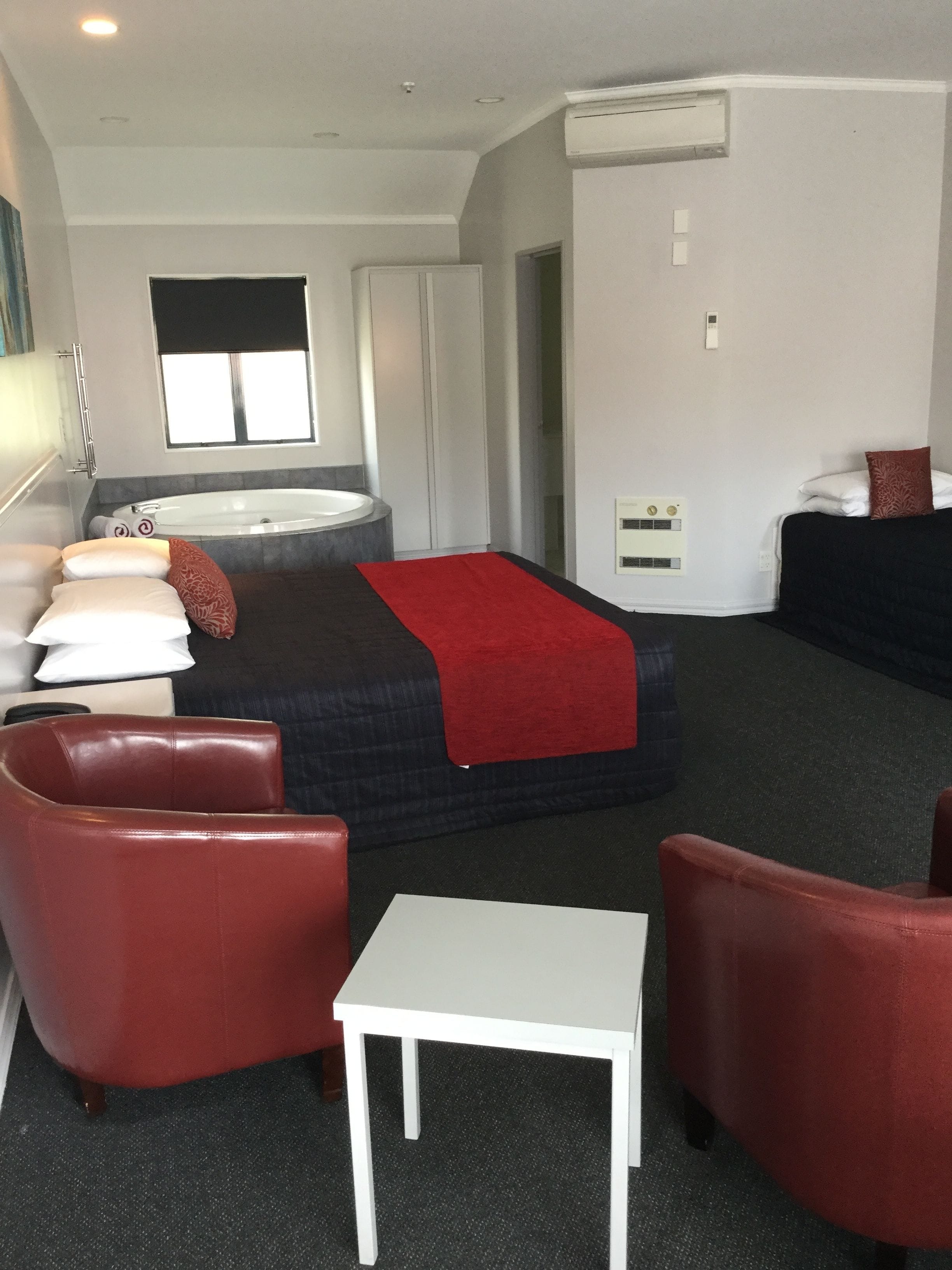 Habitación de hotel con una cama, dos sillones rojos, una mesa blanca, una bañera y un ventanal.