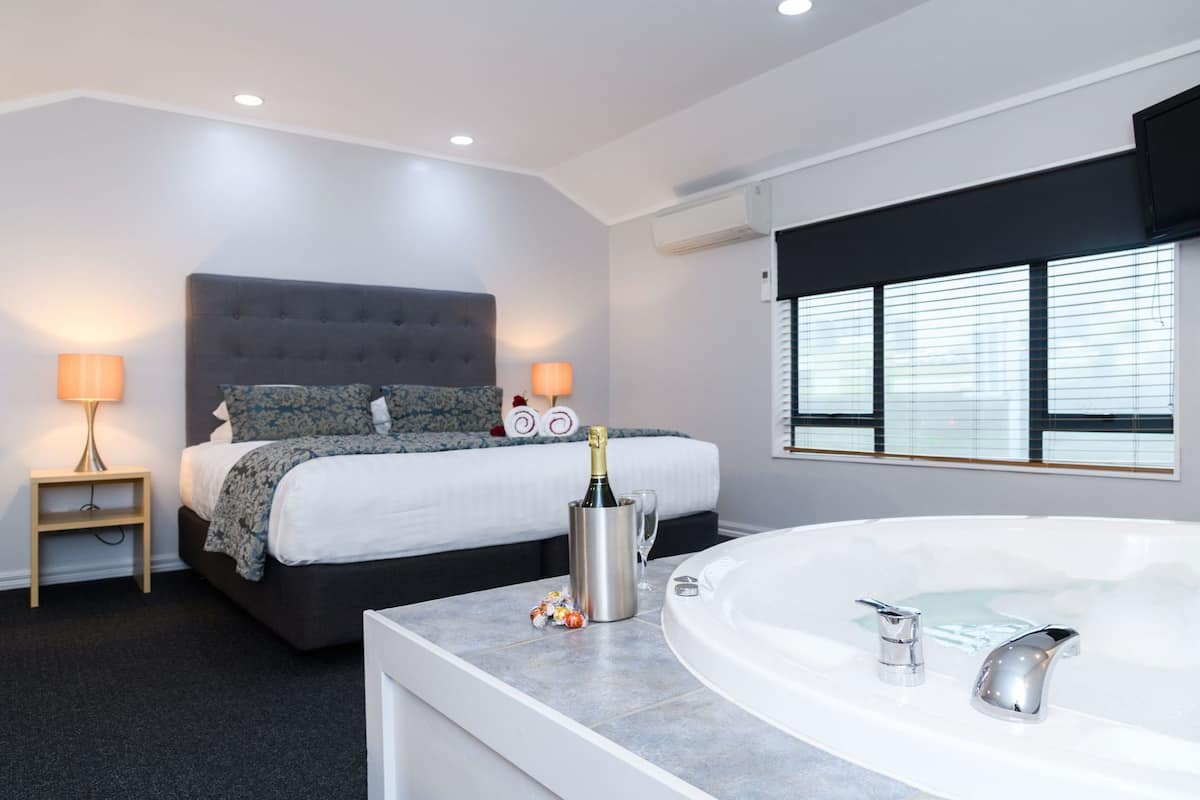 A hotel room with a bed, a sitting area with a lamp, a large bathtub, and a window with blinds.