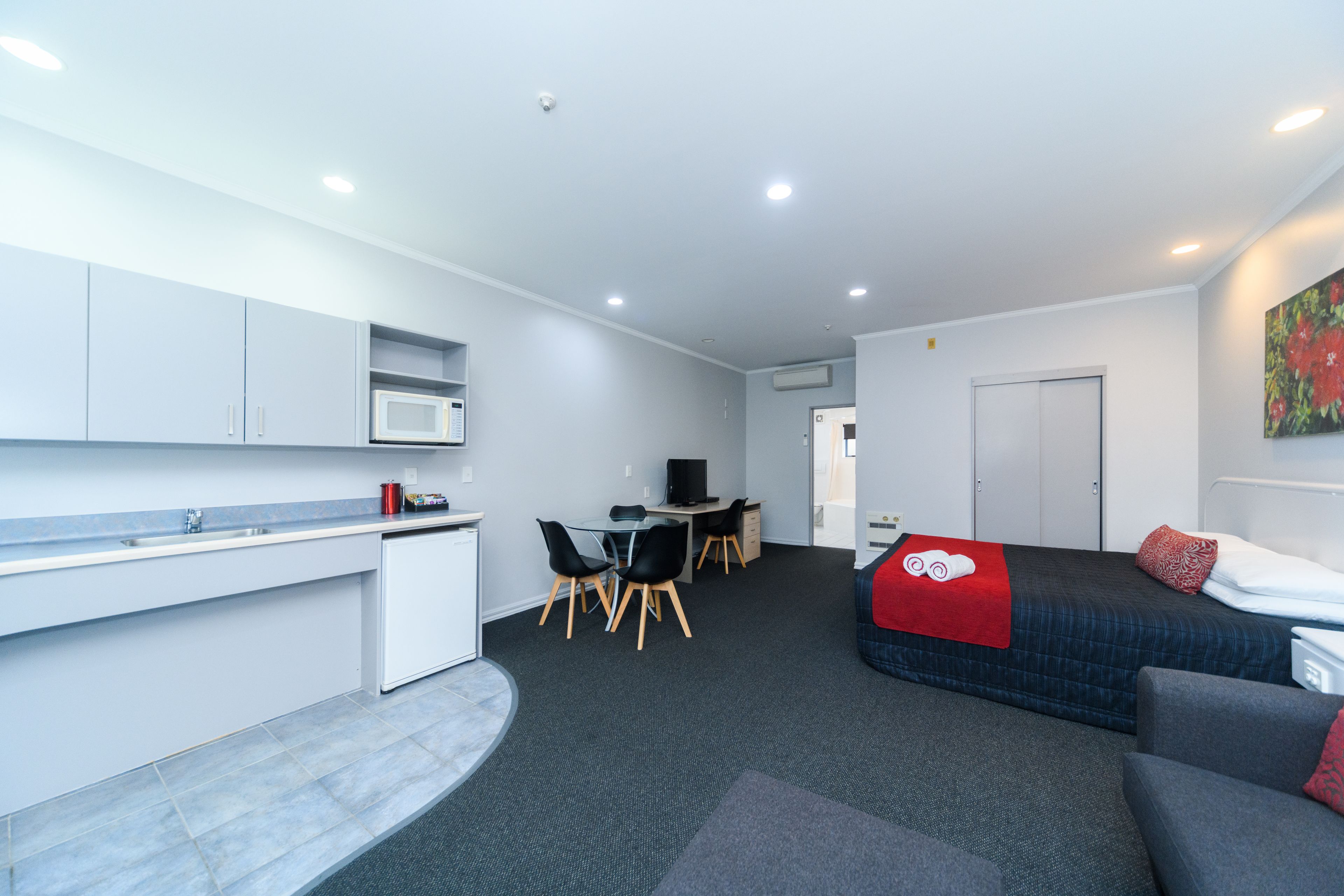 A modern hotel room with a kitchenette, a dining area, a bed with a red blanket, and a gray sofa.
