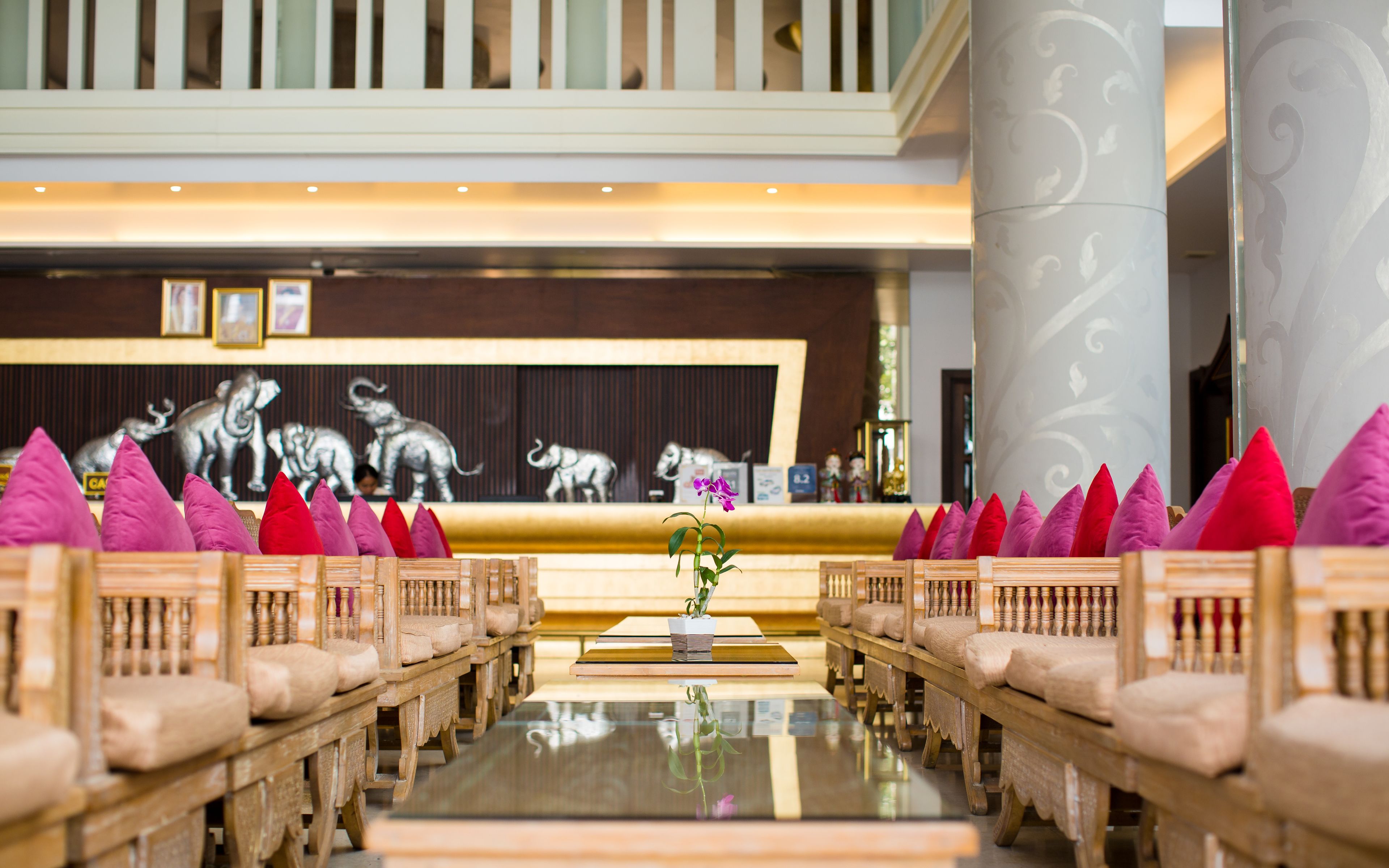 Hall d'albergo con reception, un'imponente colonna e file di poltrone in legno con cuscini.