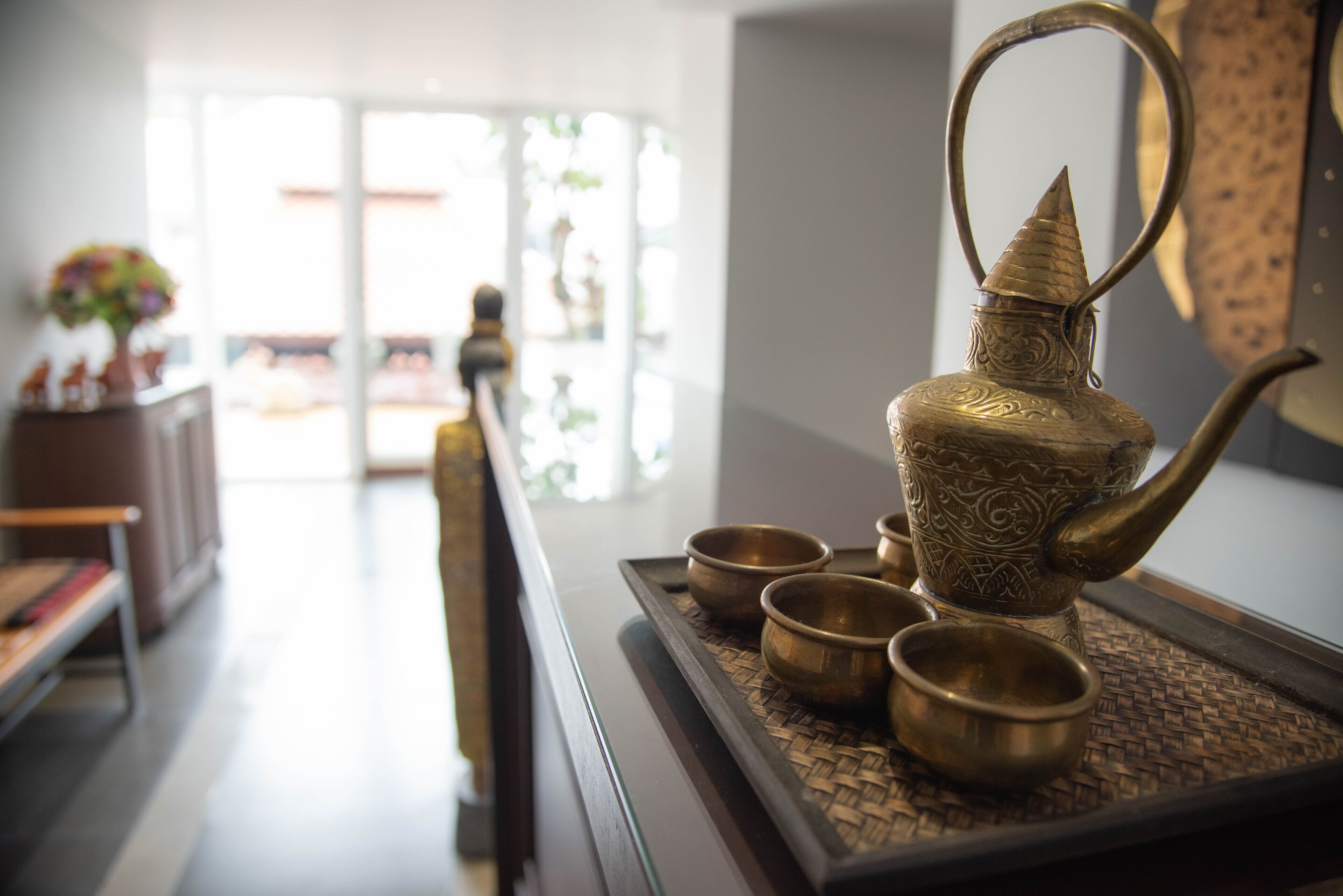 Una tetera de bronce con cuatro tazas sobre una bandeja, sobre una mesa de madera en una habitación bien iluminada con vistas a un balcón.