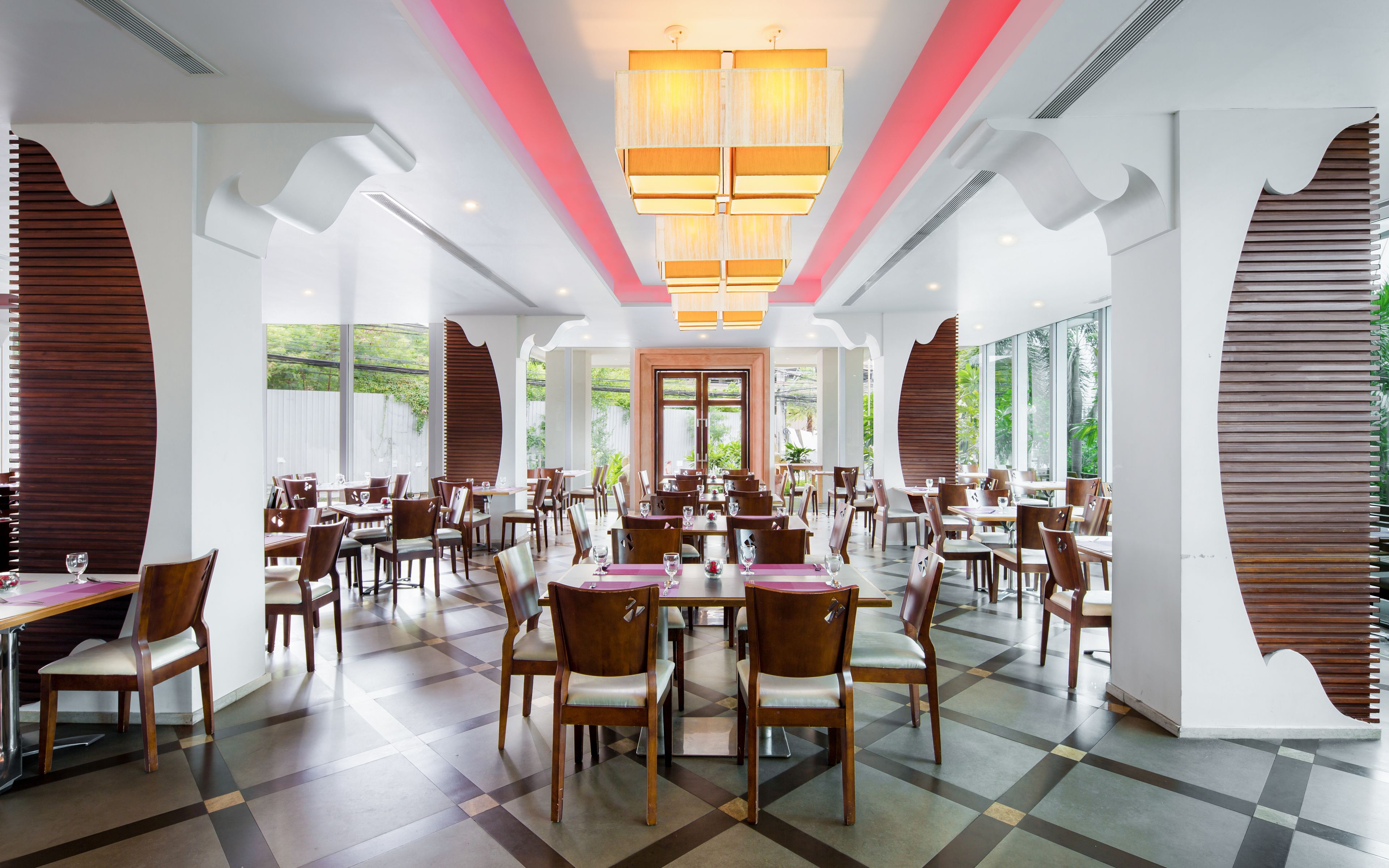 A spacious dining area with multiple tables and chairs, large windows offering views of greenery, and a modern chandelier.