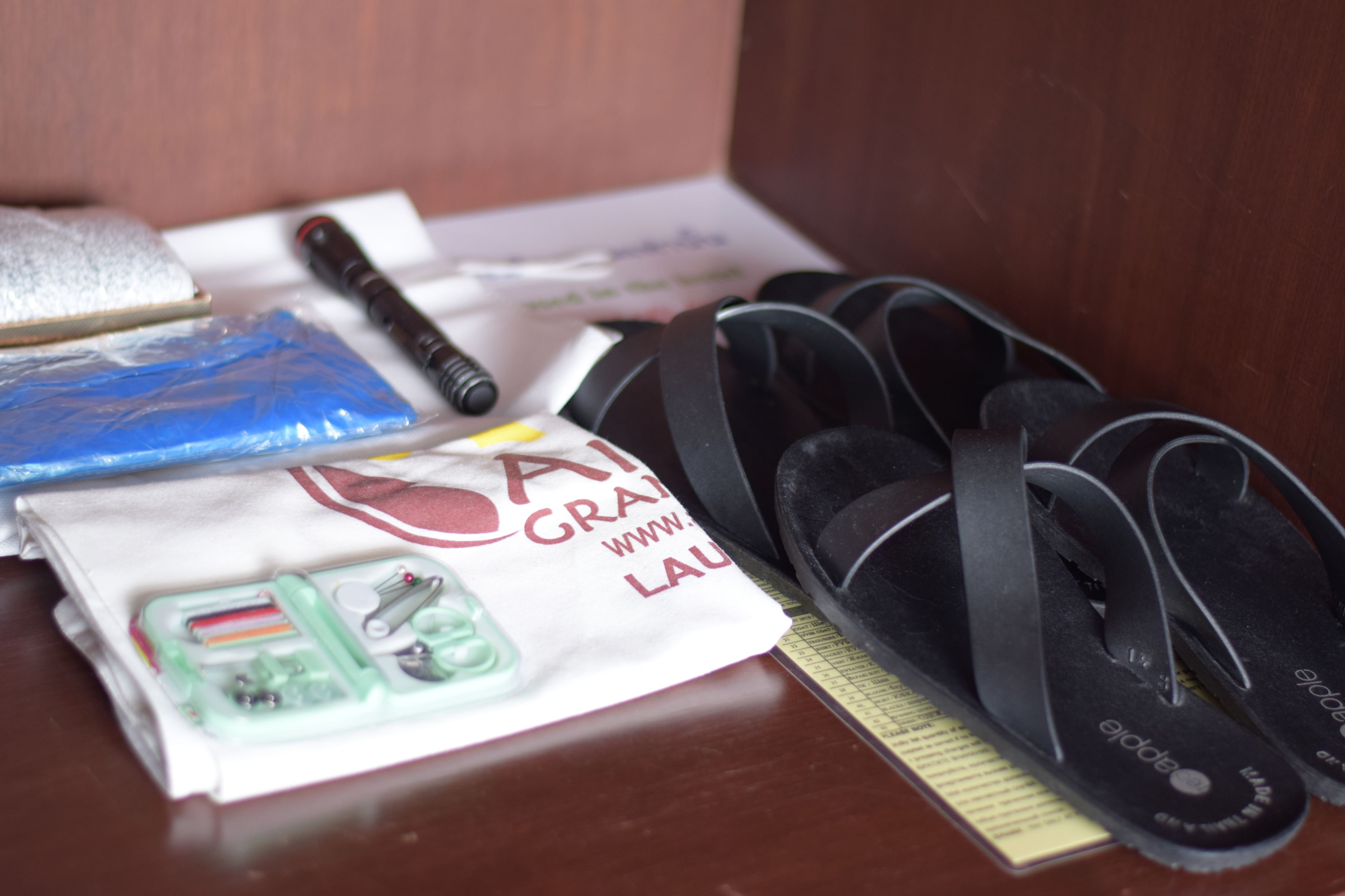 A wooden surface with a pair of black flip-flops, a white bag with red text, a blue plastic bag, and a pen.