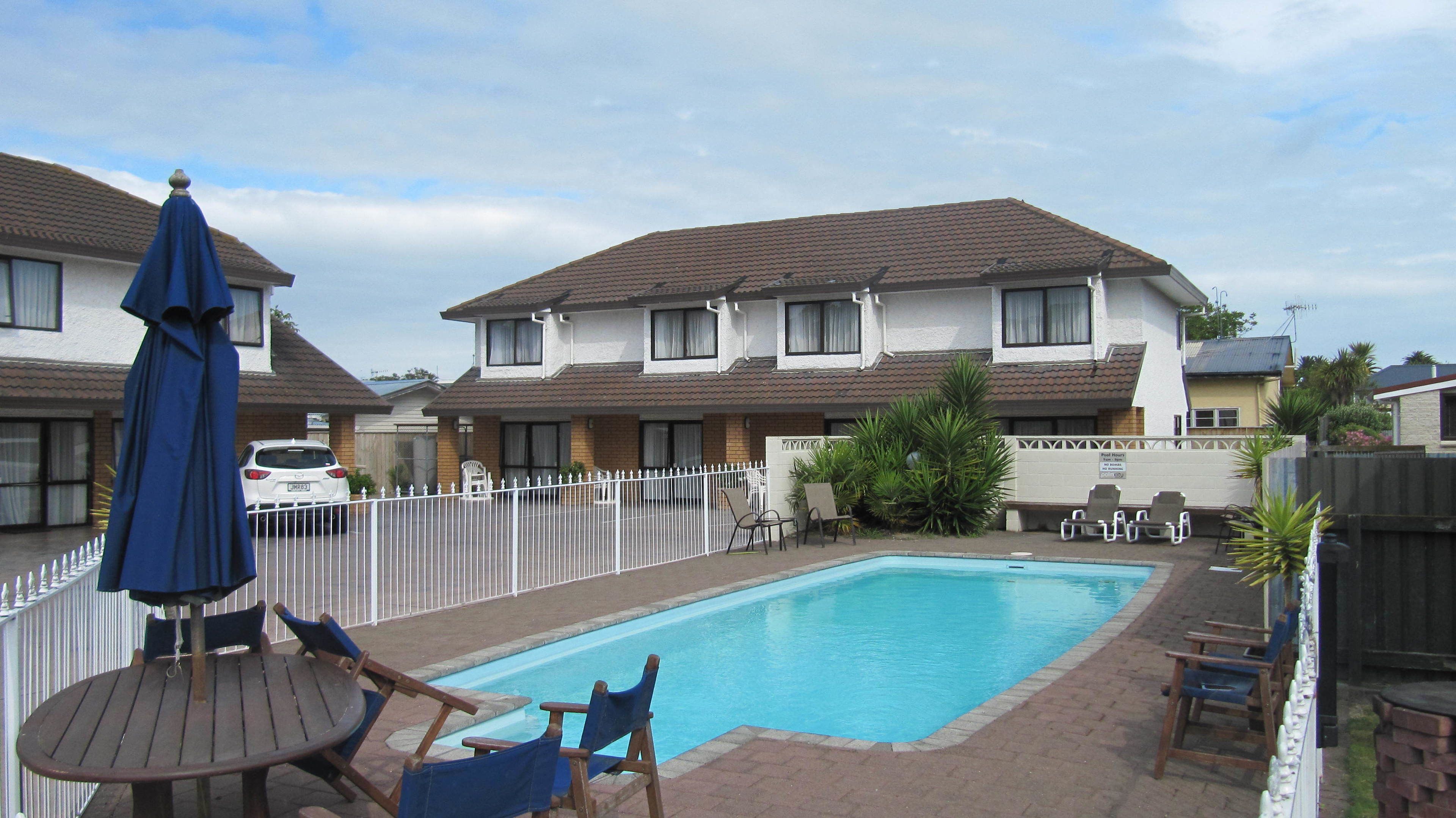 A swimming pool with lounge chairs and an umbrella, surrounded by a white fence, with a two-story building in the background.