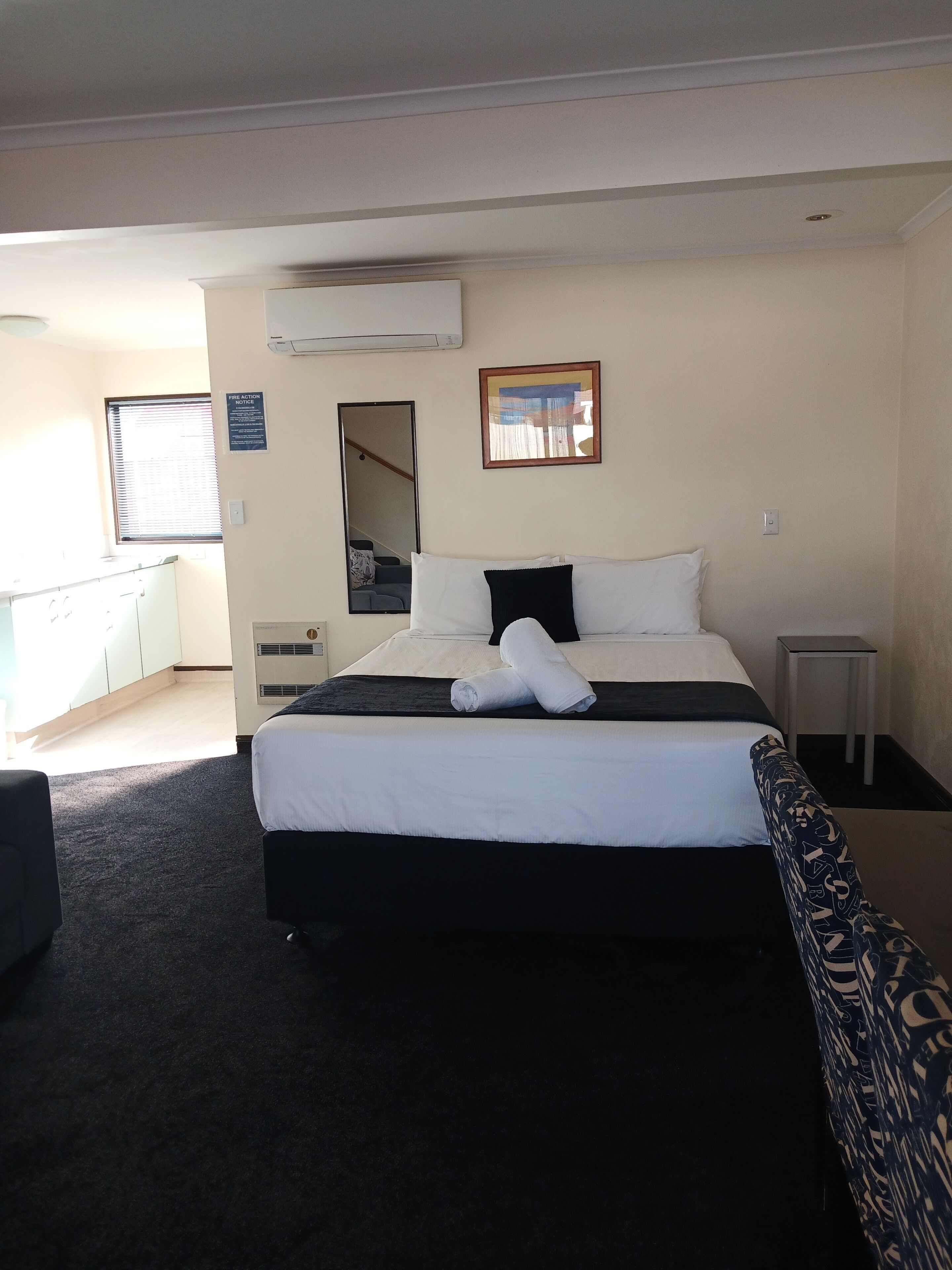 A neatly made bed with white and black bedding, a bedside table, a mirror, and a window with blinds.