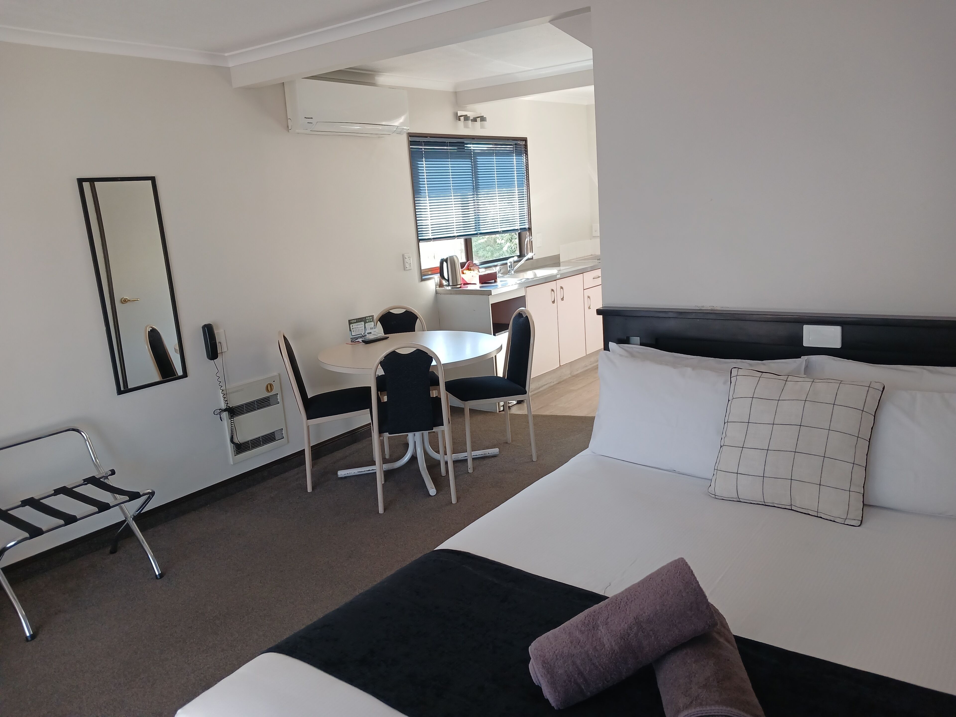 A hotel room with a bed, a desk, and a chair. There is a window with blinds, a mirror, and a heater on the wall.