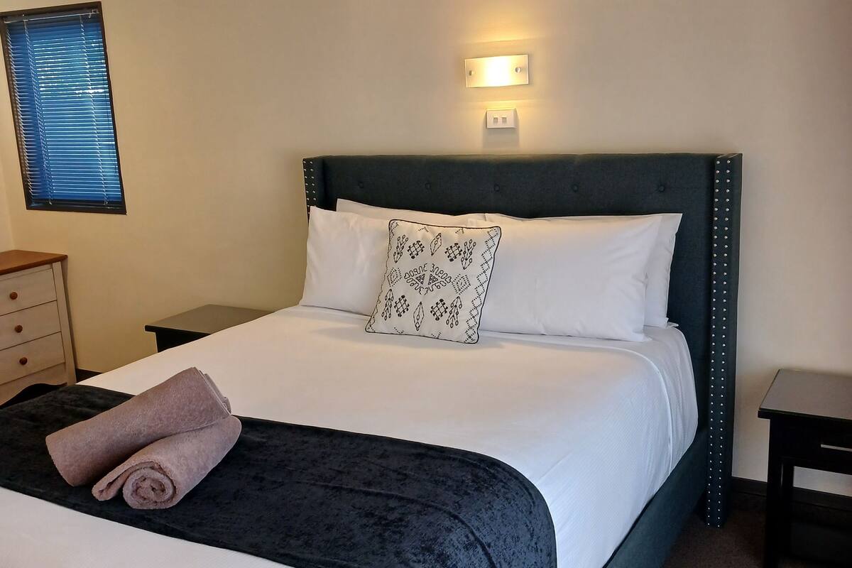 A neatly made bed with a dark headboard, white linens, and a patterned pillow. There is a small bedside table with a lamp, a window with blinds, and a wall-mounted light fixture.