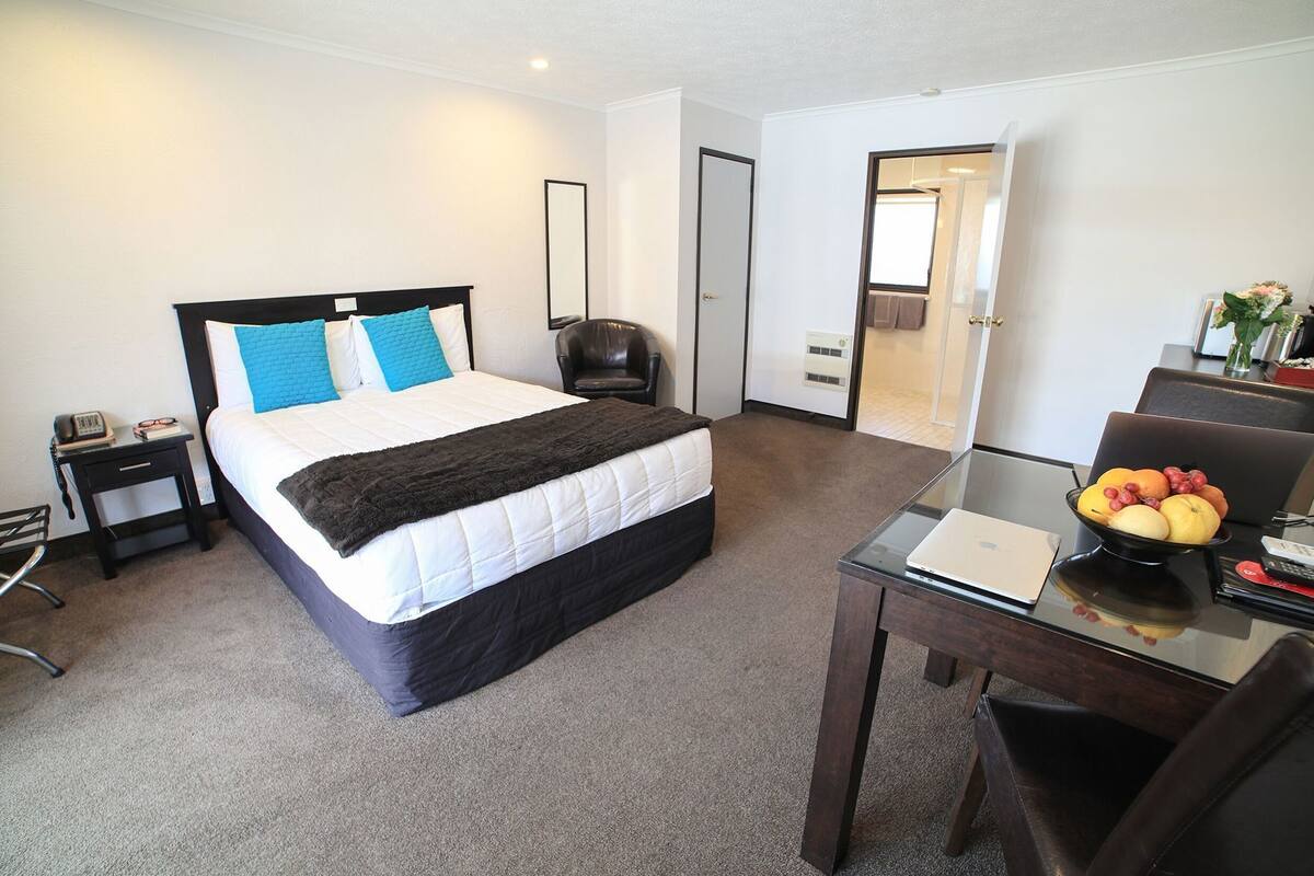 A hotel room with a bed, a desk, a chair, a mirror, a door, and a small table with fruit.