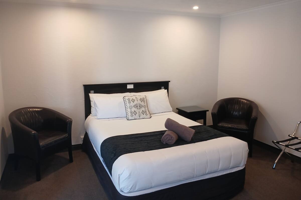A hotel room with a bed, two chairs, a small table, and a wall-mounted TV.