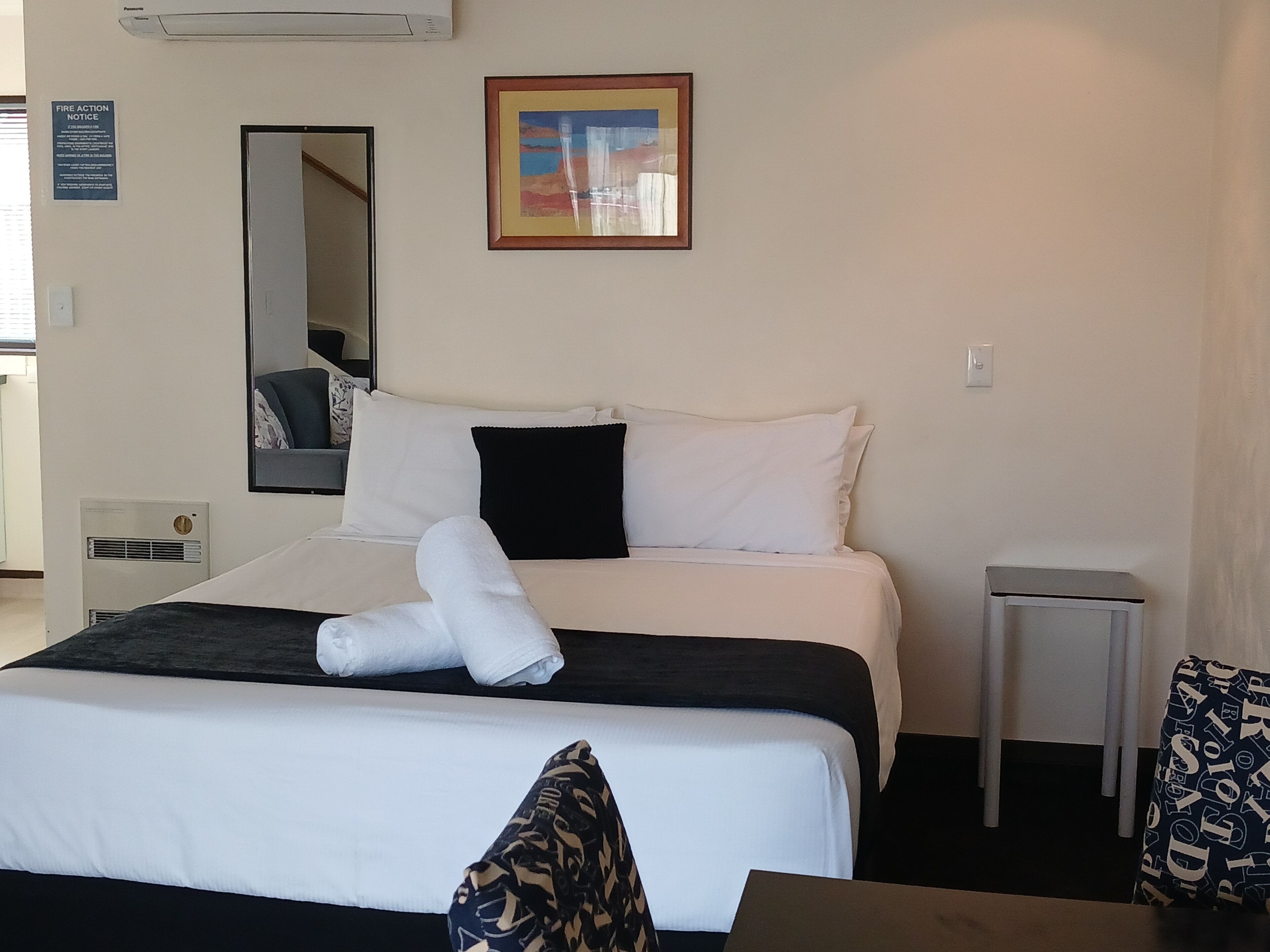 A hotel room with a bed, a bedside table, a mirror, a framed picture on the wall, and a fire action notice.