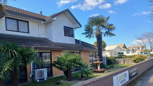 Ein zweistöckiges Haus mit Ziegelverkleidung, großen Fenstern und einem kleinen Garten mit Palmen und einer Fontäne.