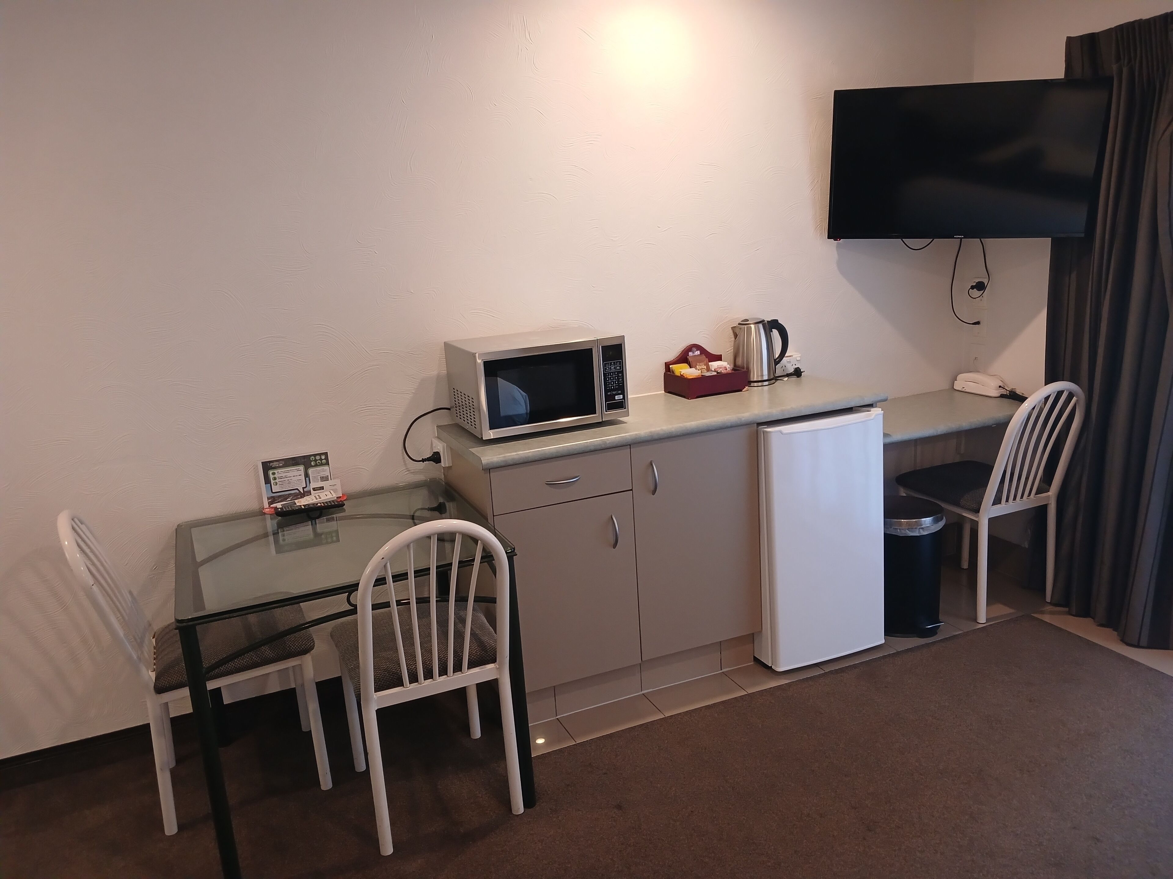 A compact kitchenette with a microwave, coffee maker, and a small refrigerator. There are two white chairs and a glass table. A TV is mounted on the wall above the kitchenette.