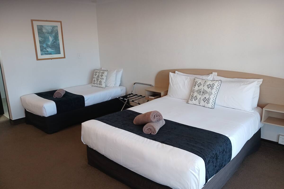 A hotel room with two single beds, each with a pillow and a rolled towel. There is a small bedside table with a lamp, a wall-mounted power outlet, and a framed picture on the wall.