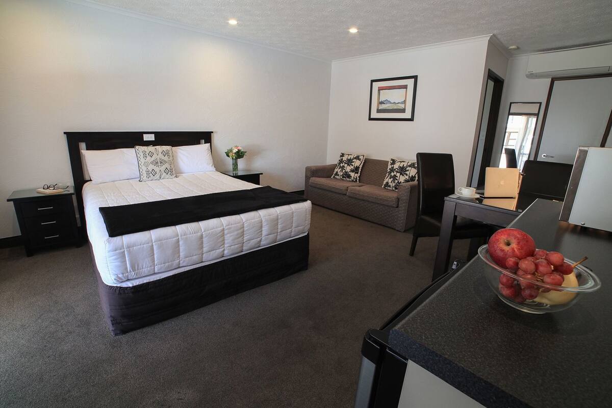 A hotel room with a bed, a sofa, a desk with a laptop, and a bowl of fruit.