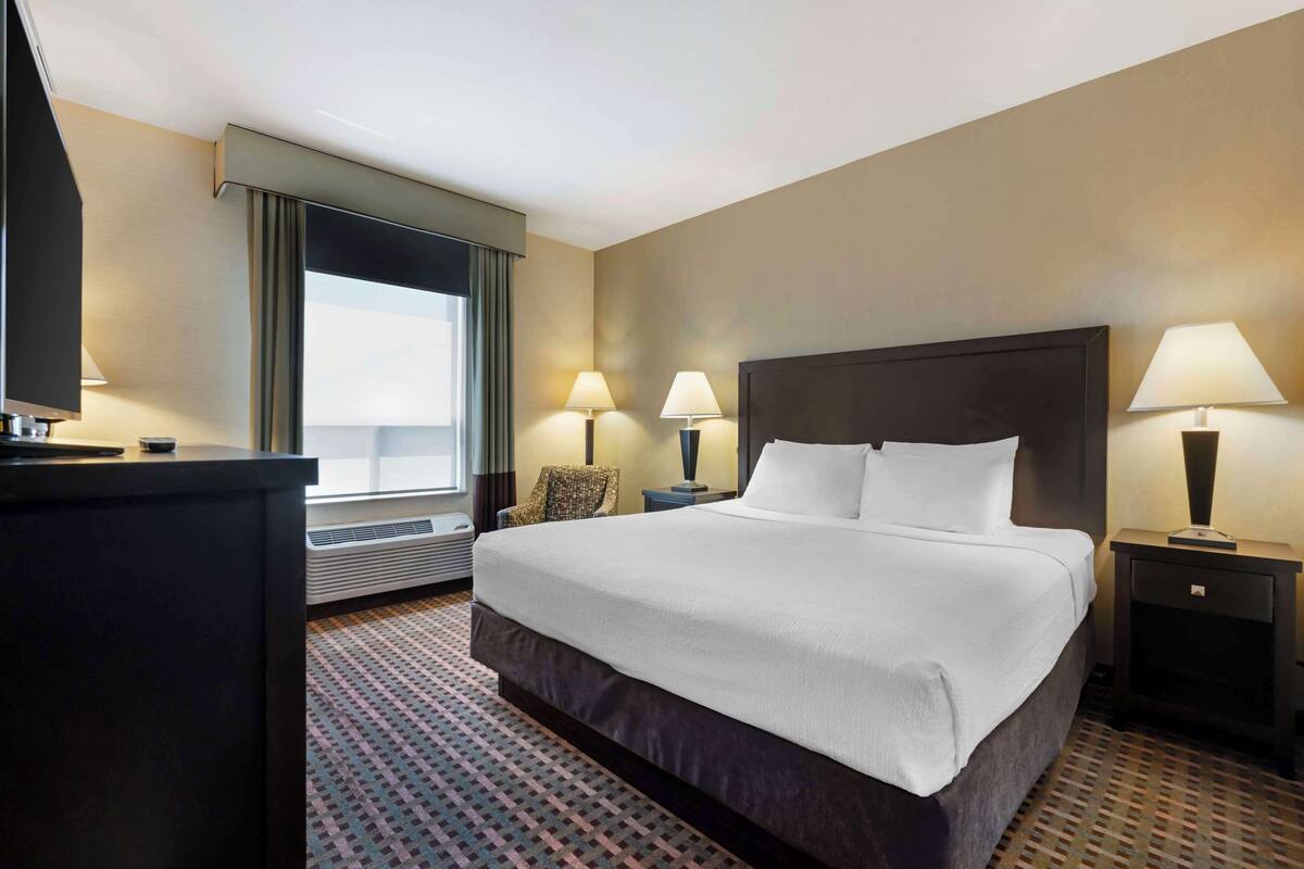 A hotel room with a large bed, two bedside lamps, a desk with a flat-screen TV, and a window with curtains.