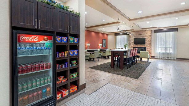 A hotel lobby with a Coca-Cola refrigerator stocked with beverages, a dining area with a table and chairs, and a flat-screen TV mounted on a stone wall. - Best Western Pacific Inn (Vernon)