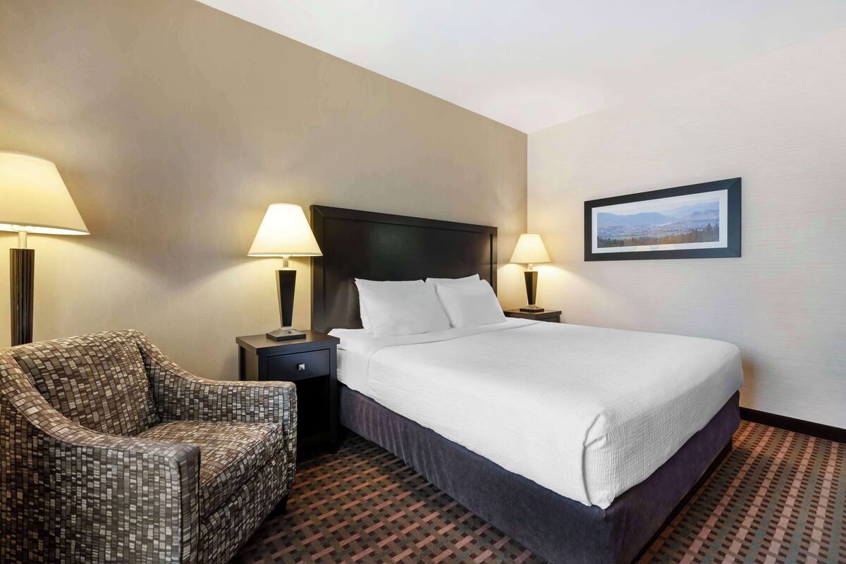 A hotel room with a large bed, two bedside tables with lamps, a patterned armchair, and a framed picture on the wall.