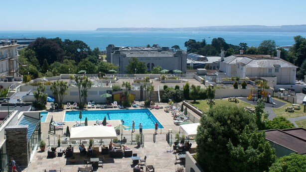 A swimming pool area with lounge chairs and umbrellas, surrounded by buildings and trees.