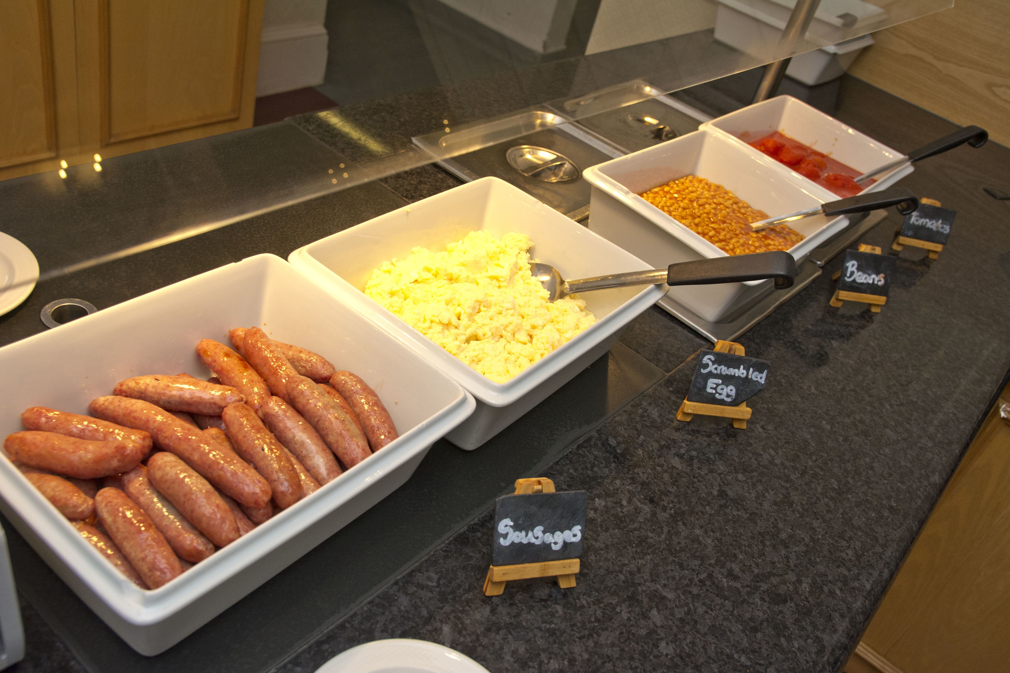 A buffet with labeled containers of sausages, scrambled eggs, and beans.