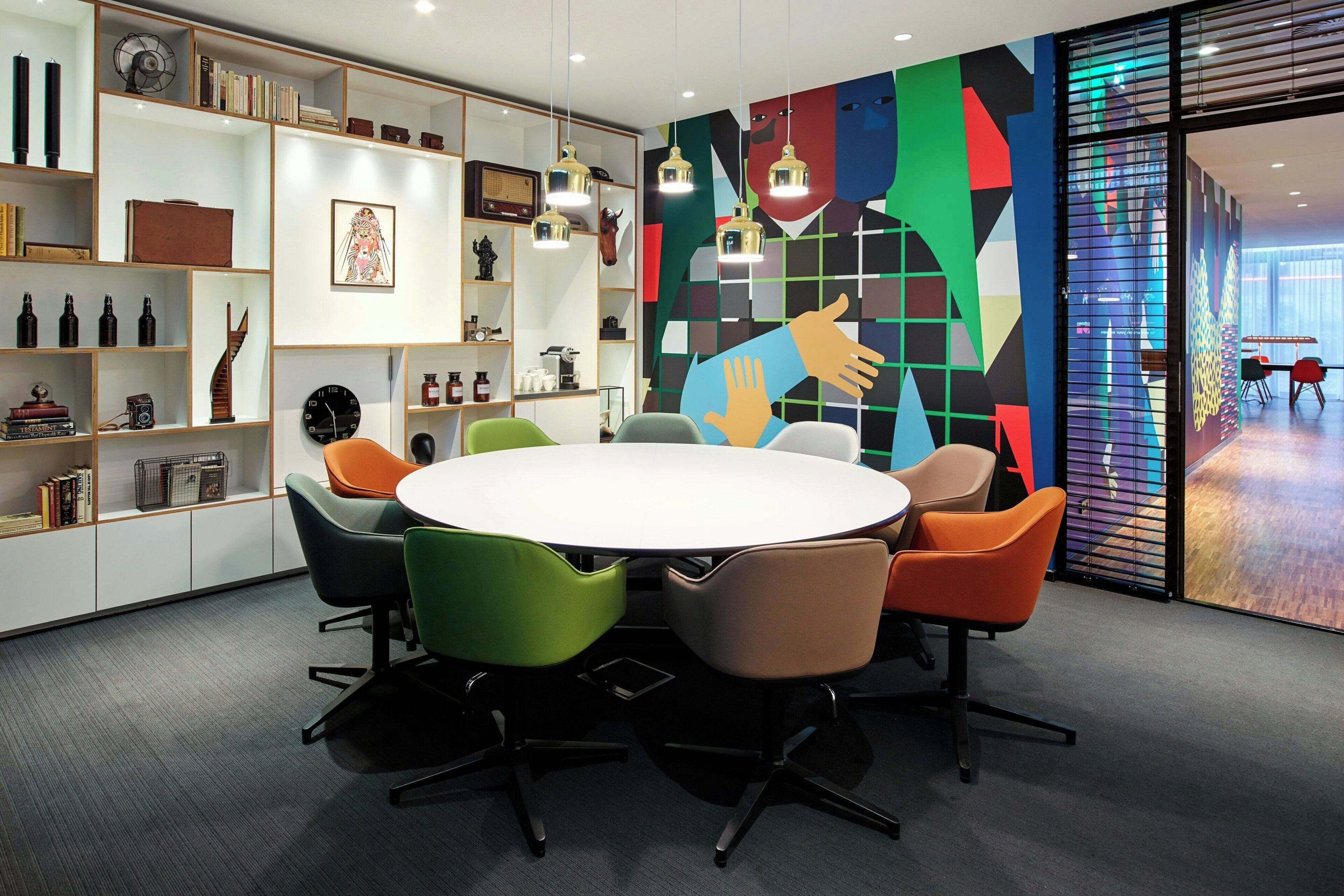 A modern meeting room with a round table, colorful chairs, and a vibrant abstract mural.