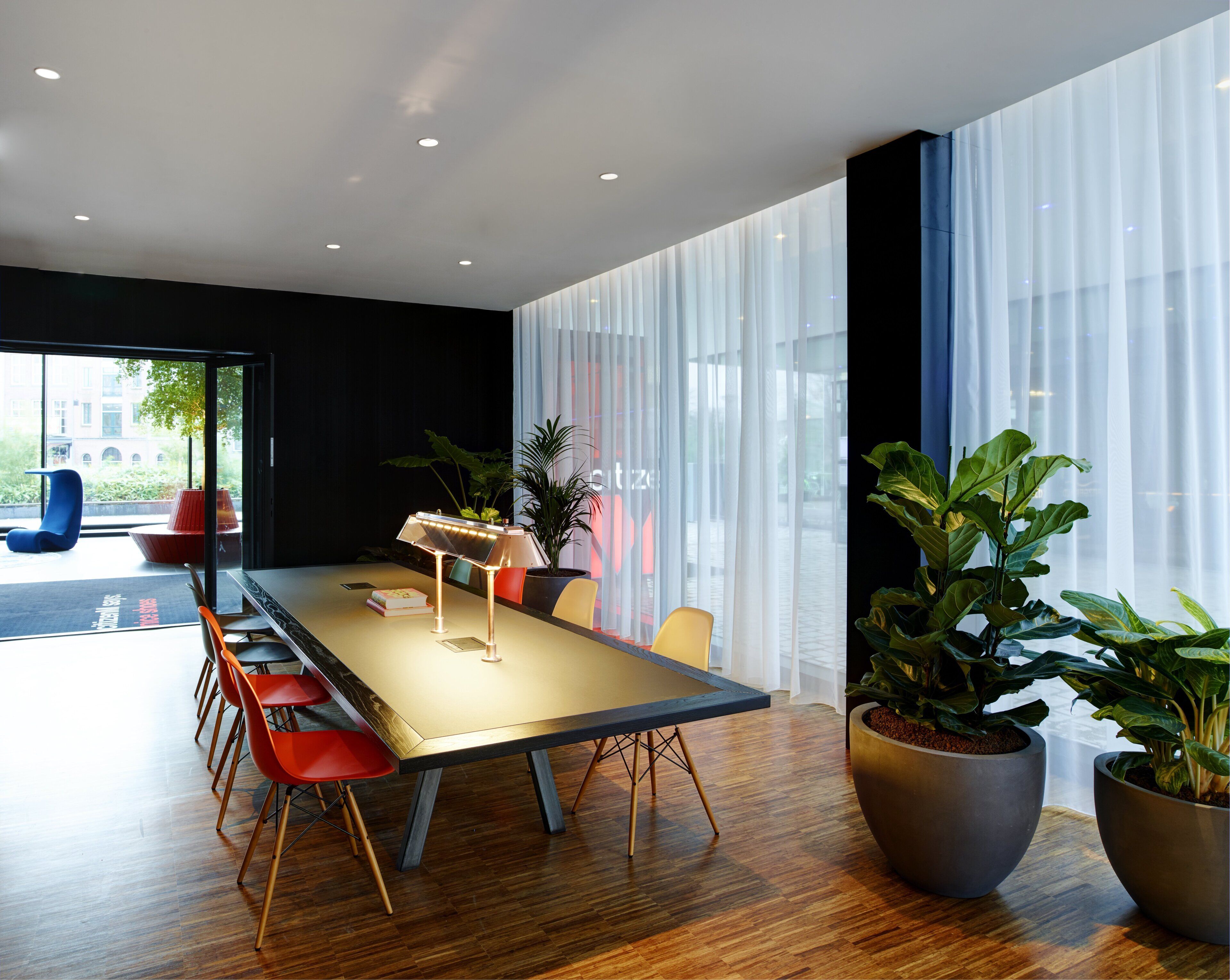 A modern meeting room with a long table, several chairs, potted plants, and large windows with sheer curtains.