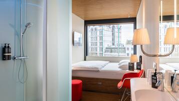 Een moderne hotelkamer met een douche, een bed, een rode stoel, een groot raam en een badkamer met een wastafel en spiegel.