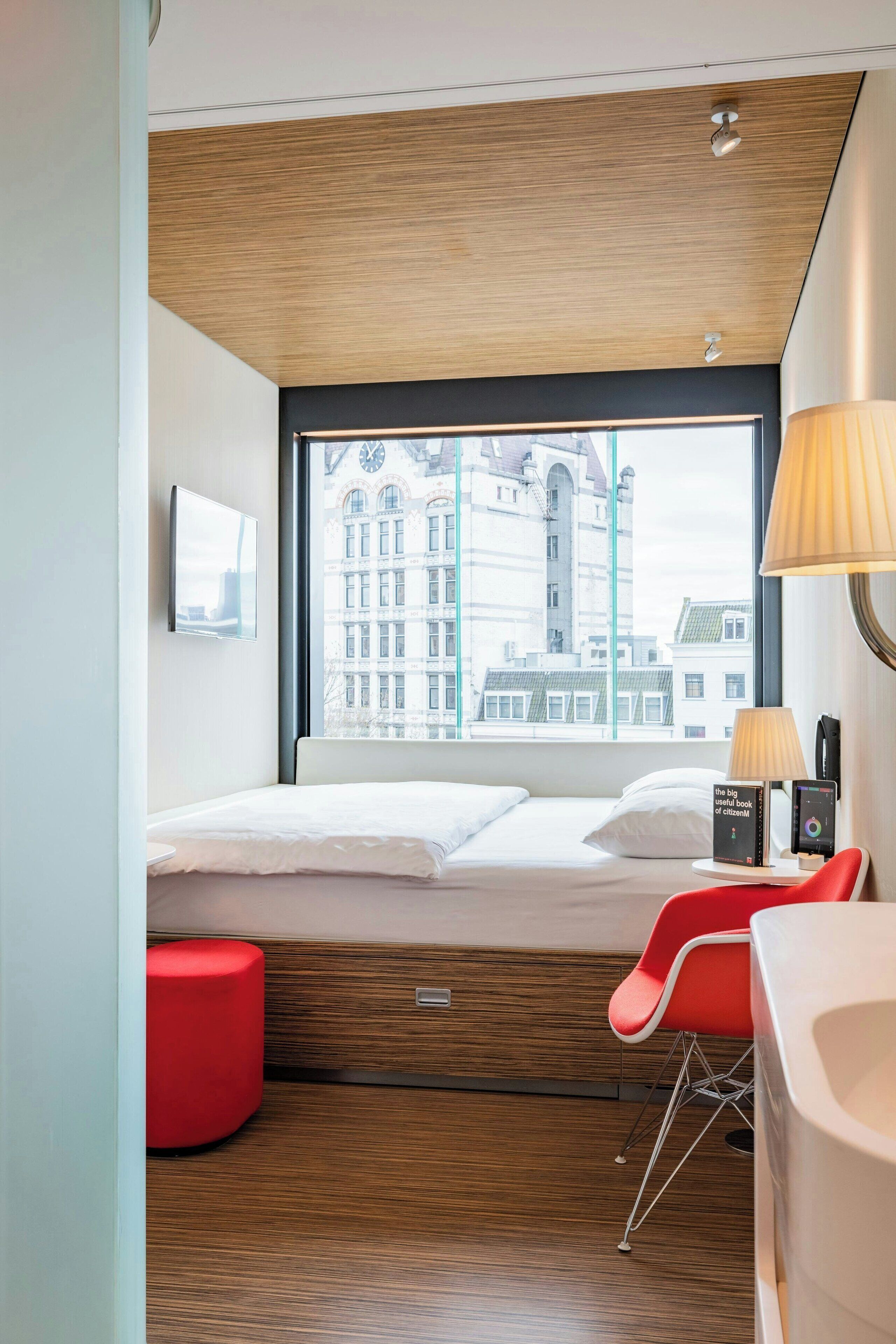 A modern hotel room with a large window, a bed, a red chair, and a desk with a lamp.