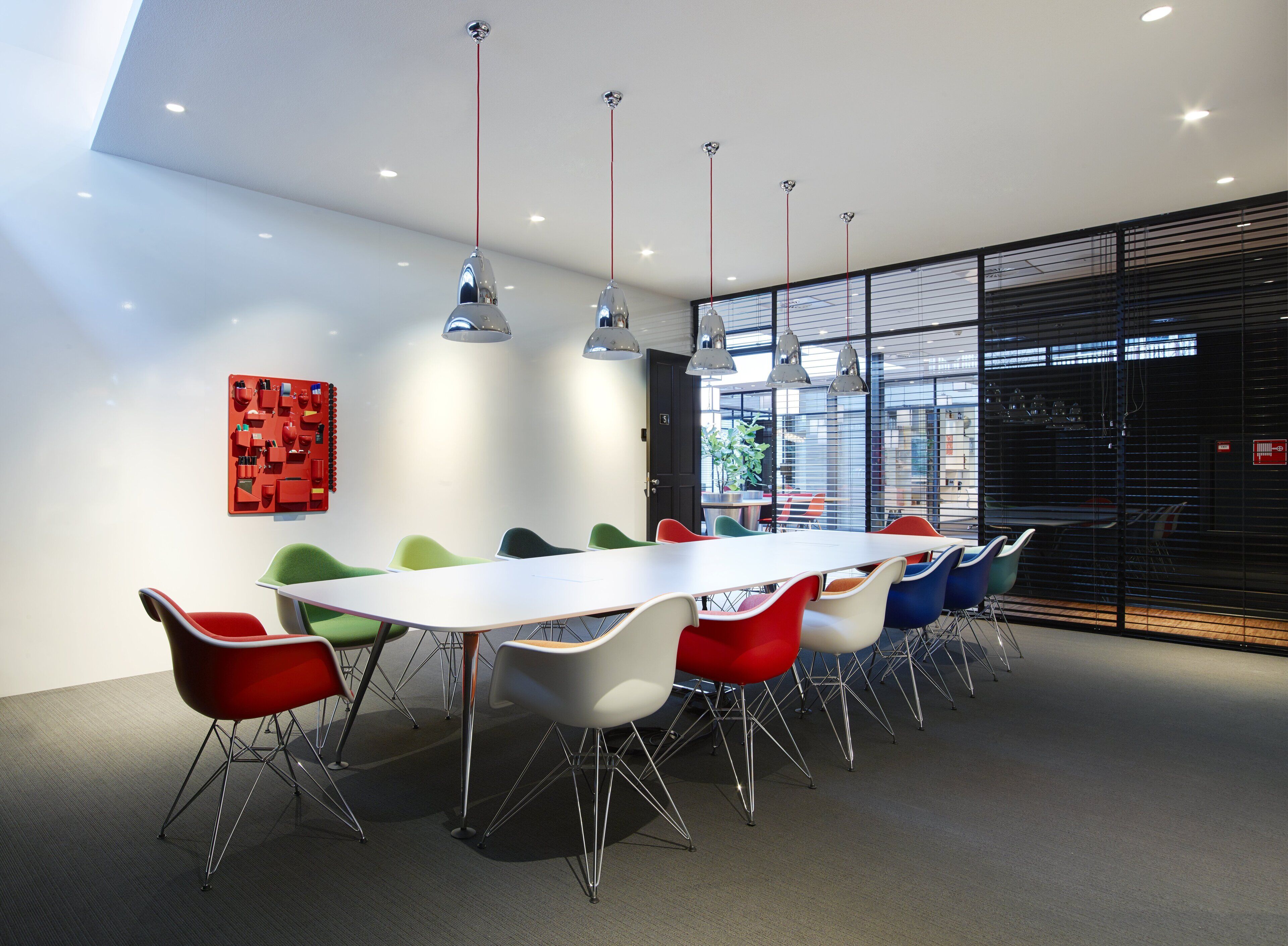 Une salle de conférence moderne, dotée d’une longue table, de chaises colorées, de luminaires suspendus et d’une grande fenêtre avec des stores.