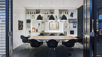 Una sala de conferències moderna amb una taula de fusta, cadires negres, llums penjants i una biblioteca amb diversos objectes.