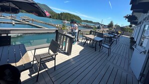 A wooden deck with outdoor seating, a view of a marina with boats, and a mountain in the background.