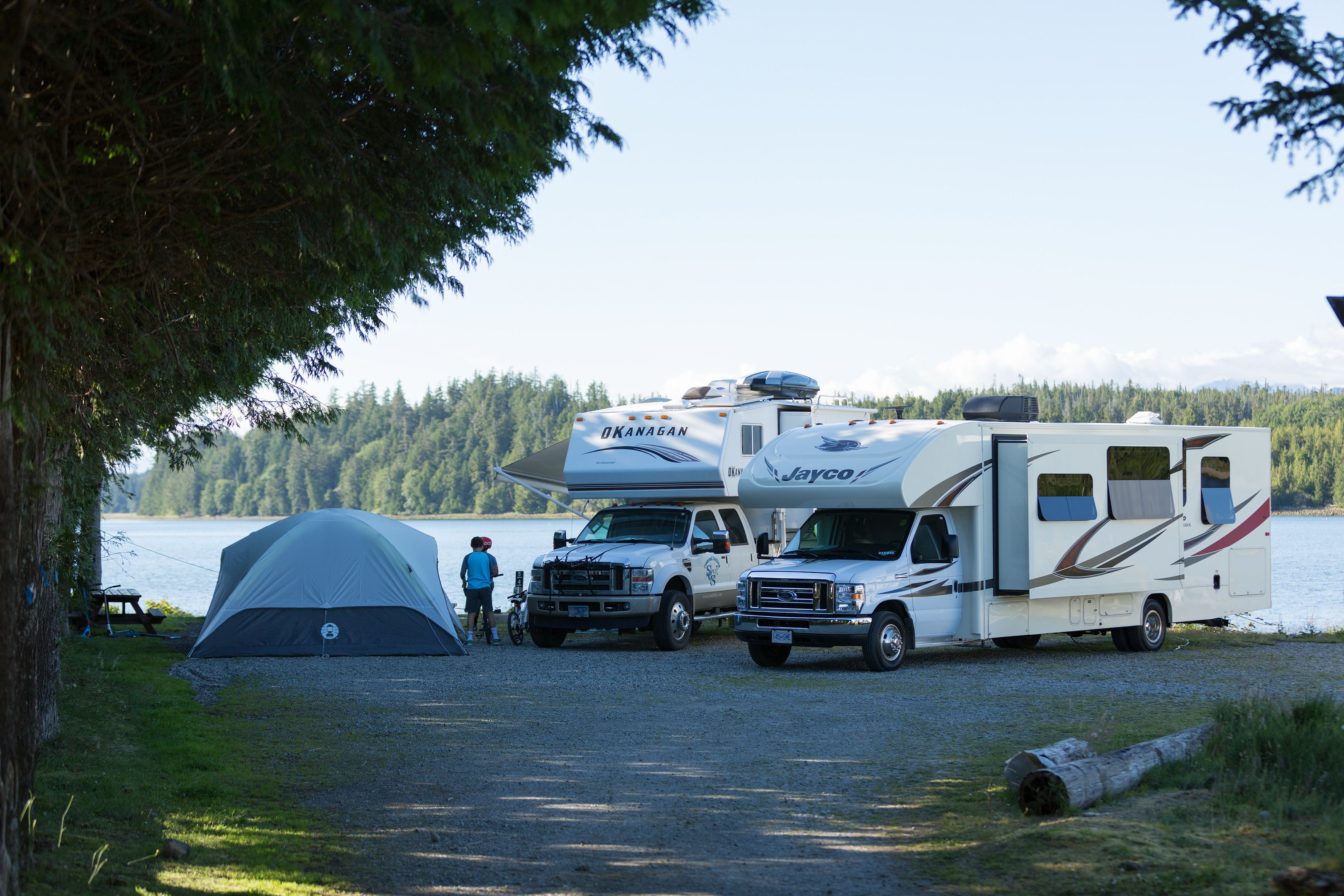 Ein Campingplatz mit Zelt, zwei Wohnmobilen und einem geparkten Lastwagen am See.
