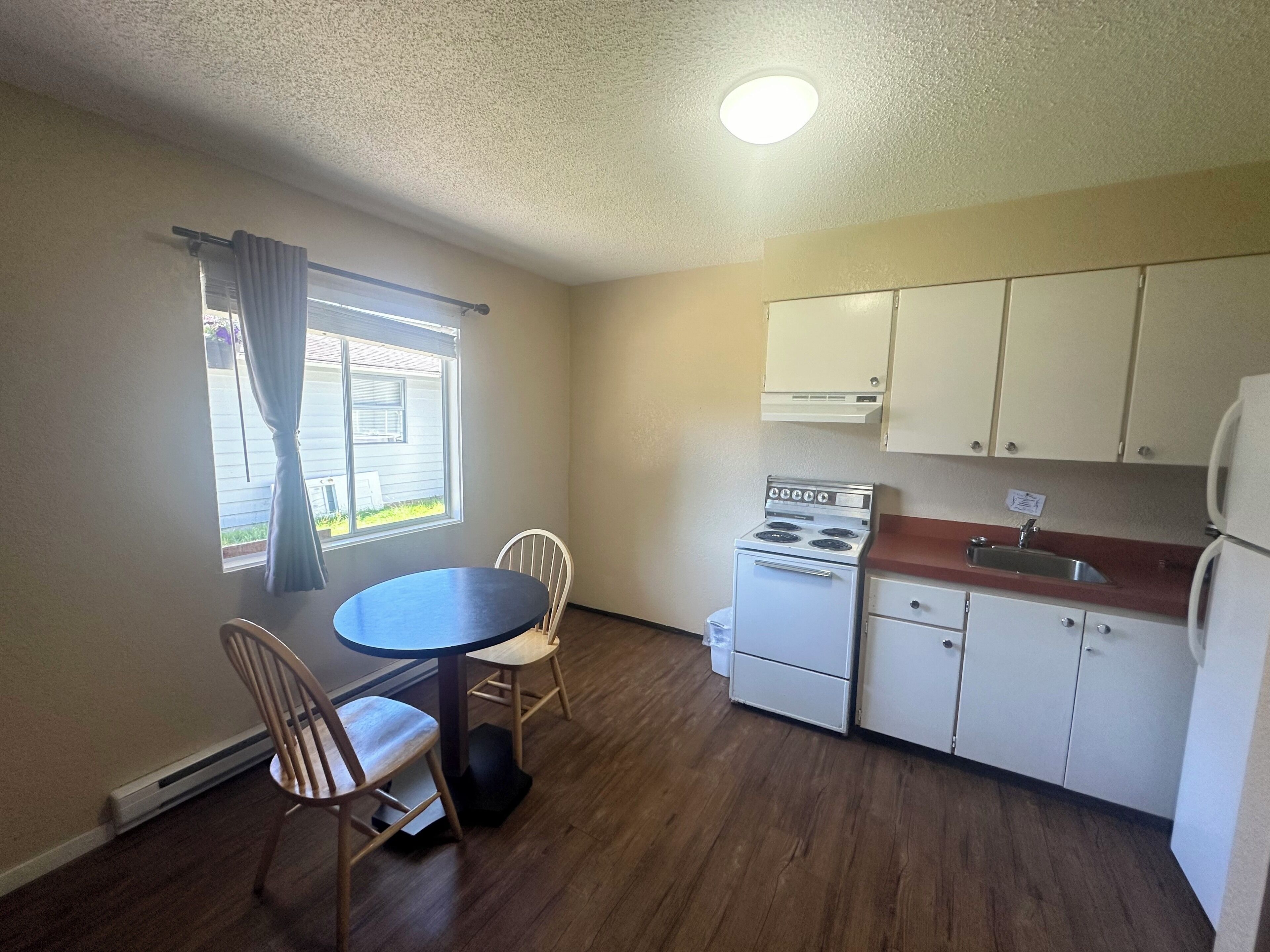 A compact kitchen with a stove, sink, and cabinets. There is a dining table with chairs and a window with curtains.