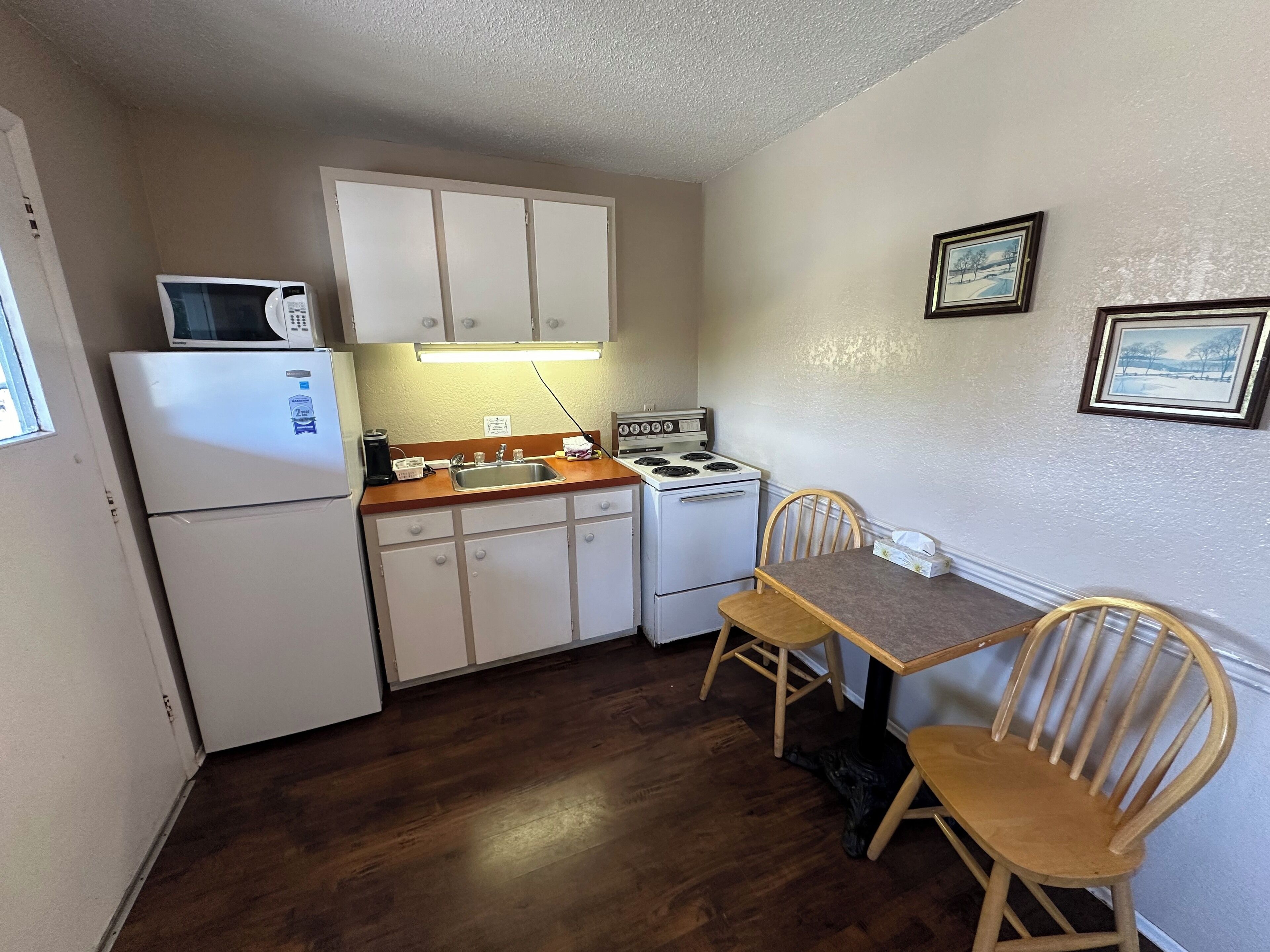 A compact kitchen with white cabinets, a microwave, a stove, and a sink. There is a wooden table with two chairs and a cat on the floor.