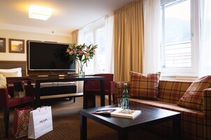 Habitación de hotel con sofá, una mesa de centro con un libro y vasos, televisor, jarrón con flores y ventana con cortinas.
