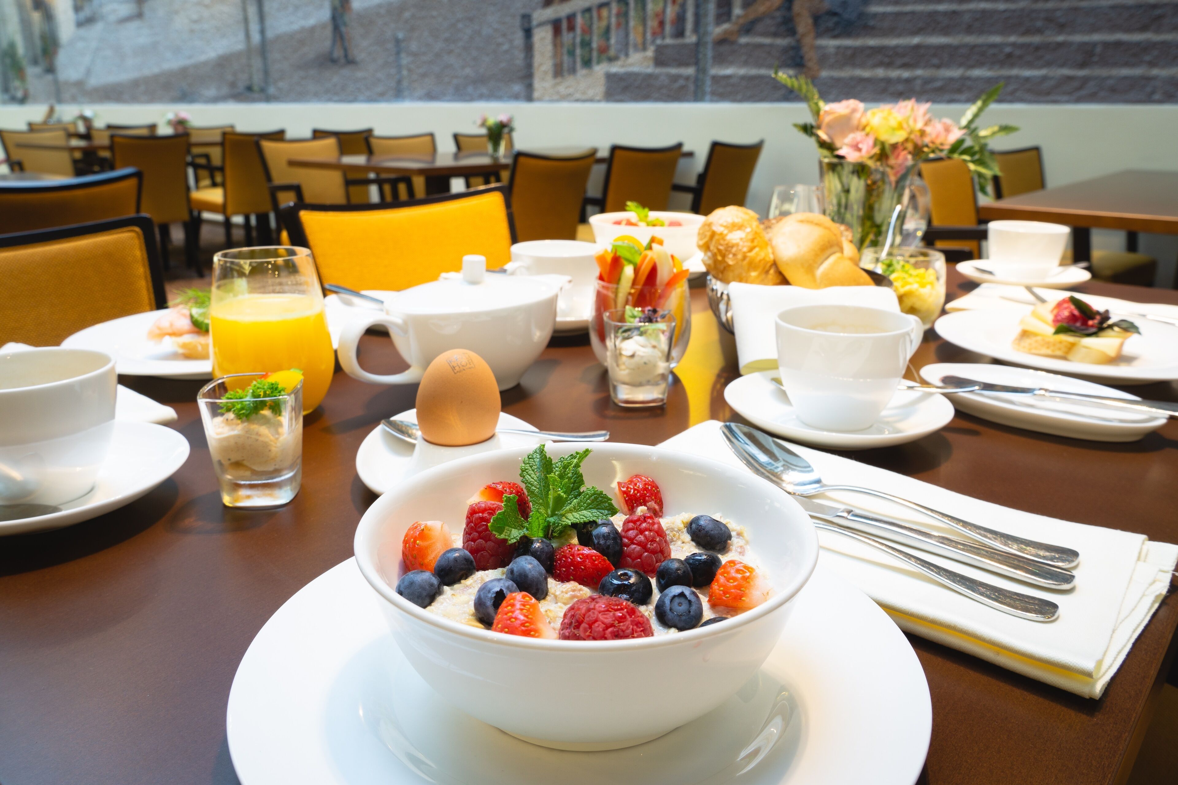 Desayuno con un bol de fruta, un vaso de zumo de naranja y diferentes alimentos sobre una mesa.