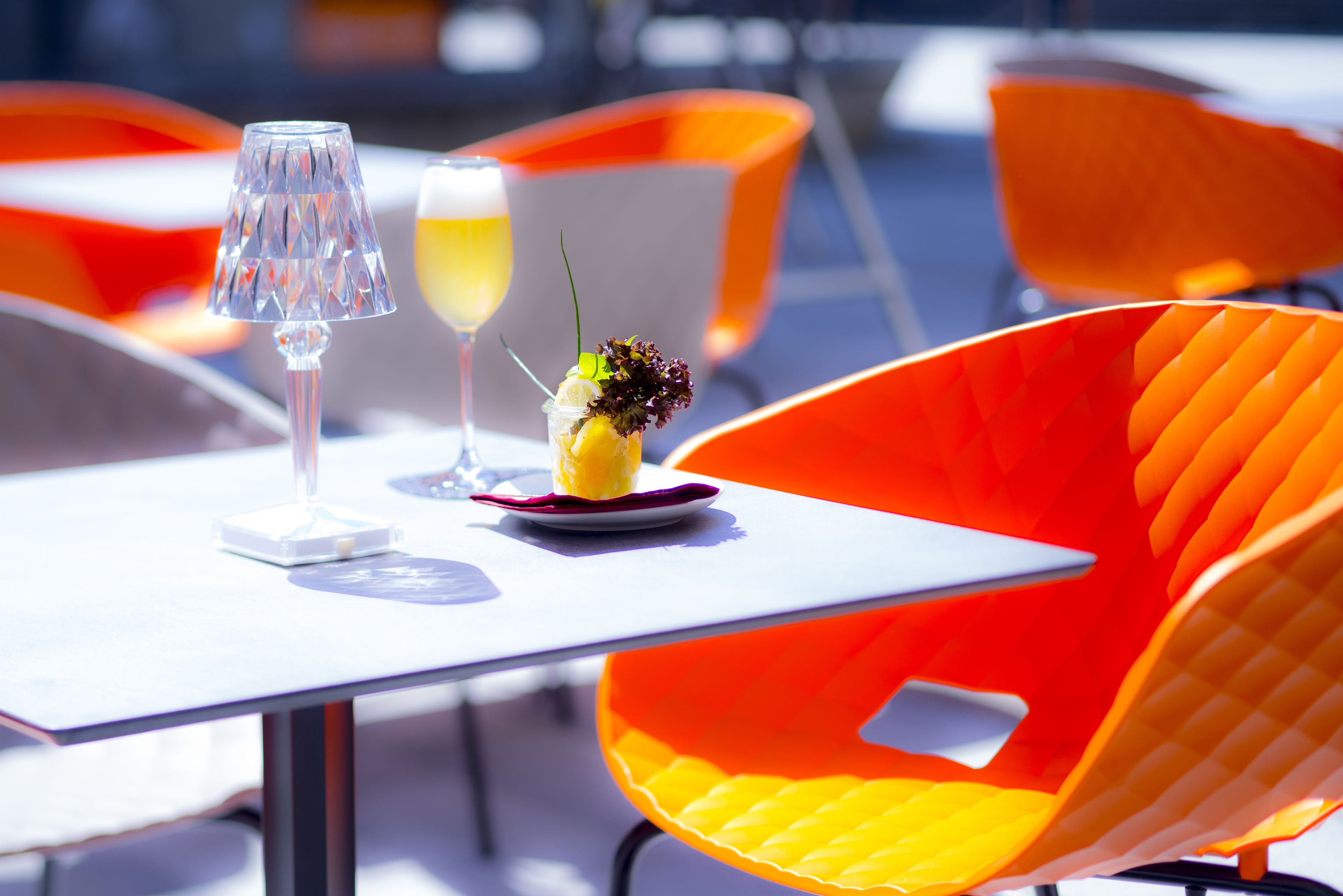 Una mesa con una copa y una bebida, un arreglo floral y sillas color naranja.