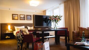Une chambre d’hôtel avec un lit, une télévision, une table à manger, un canapé rouge, un fauteuil rouge, un vase avec des fleurs et une fenêtre avec des rideaux.