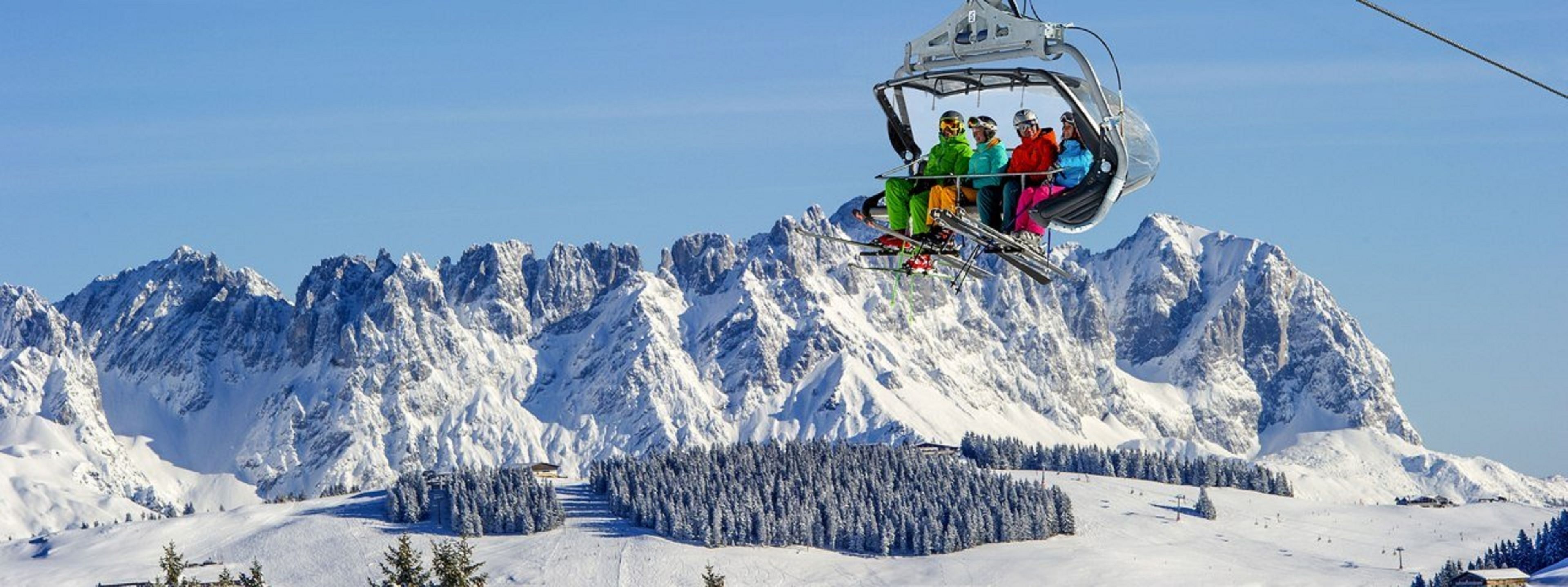 A ski lift with passengers, snow-covered mountains, and a forested area in the background.