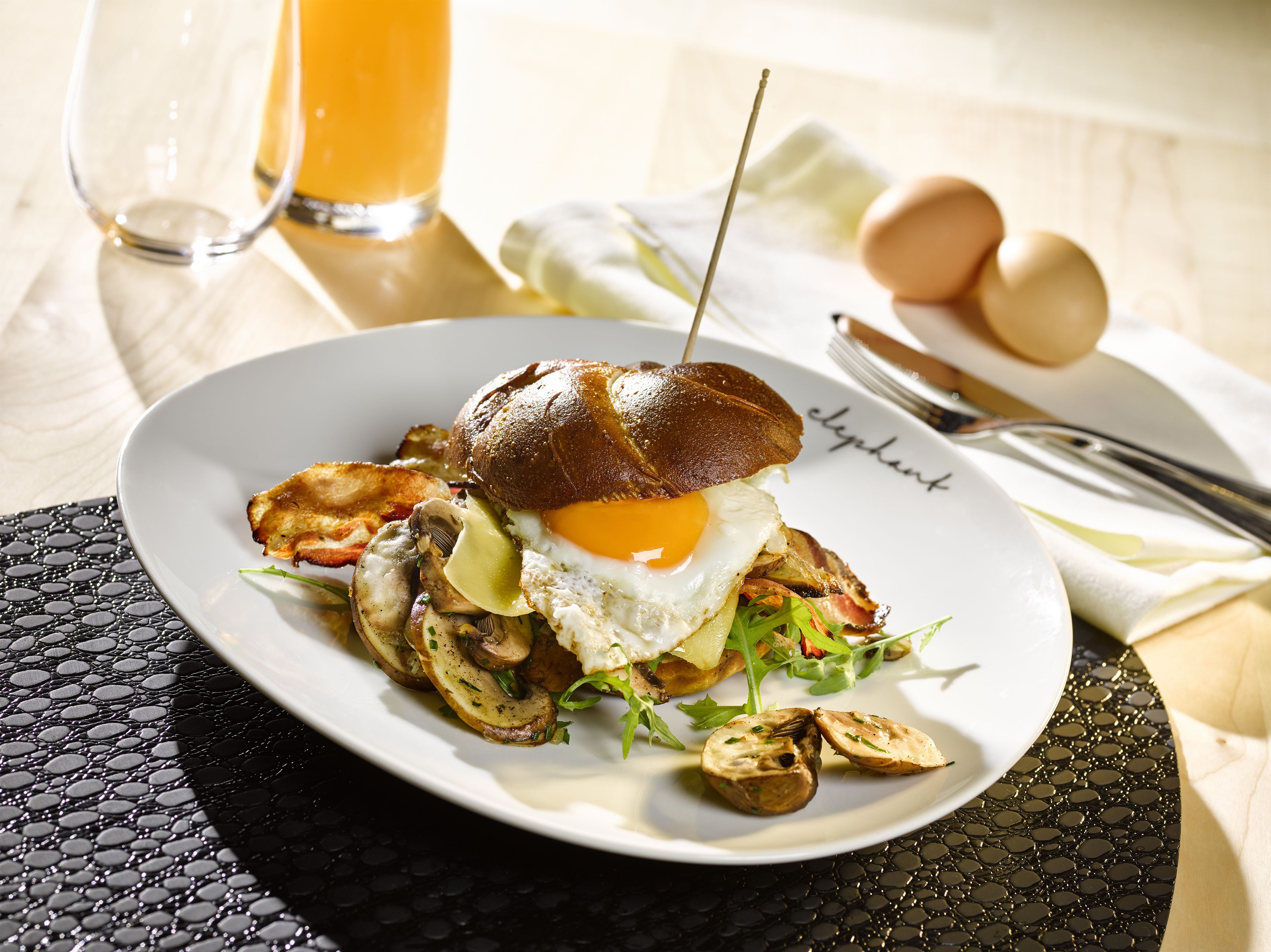 Un plato con un sándwich, huevos, champiñones, verduras de hoja y un vaso de zumo de naranja, además de una servilleta con dibujos de huevos sobre una mesa.