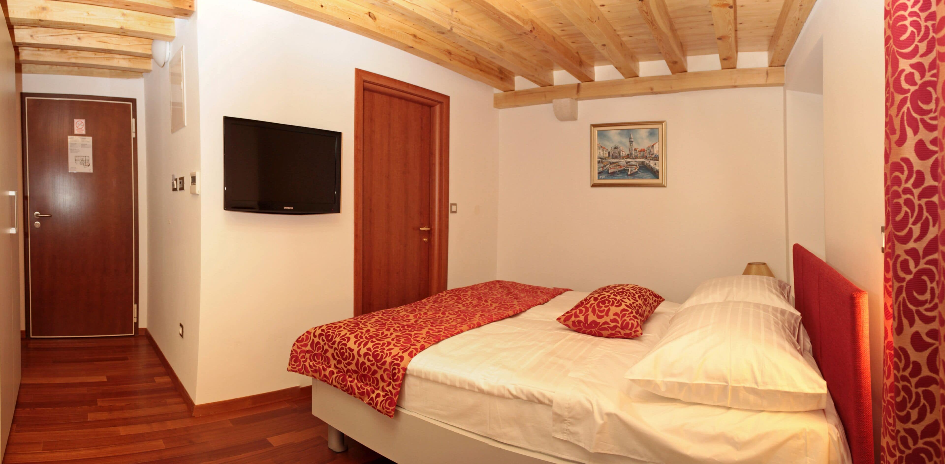 A bedroom with a wooden ceiling, a bed with red and white bedding, a television on the wall, and a painting above the bed.