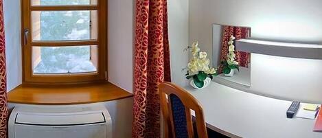 A corner room with a window, a chair, a small table with a vase of flowers, and a white wall with a mirror.