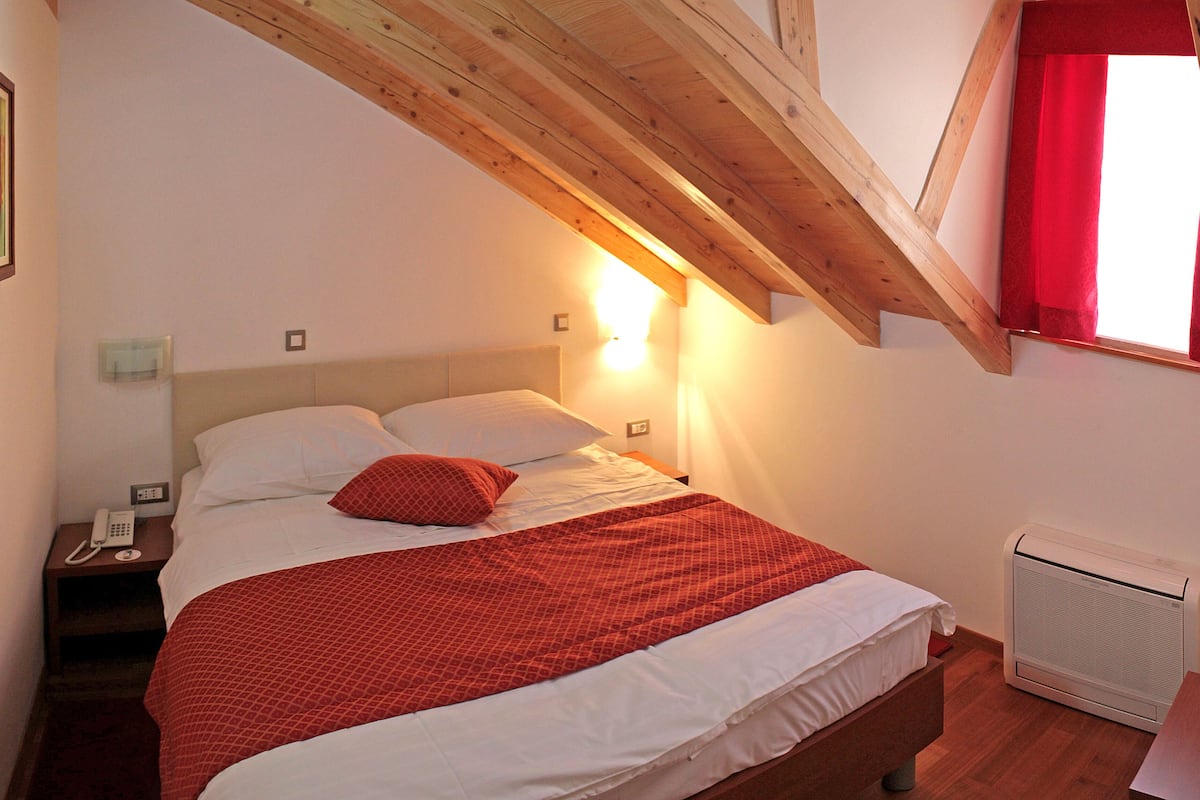 A bedroom with a wooden ceiling, a bed with white bedding and a red blanket, a bedside table with a phone, and a radiator.