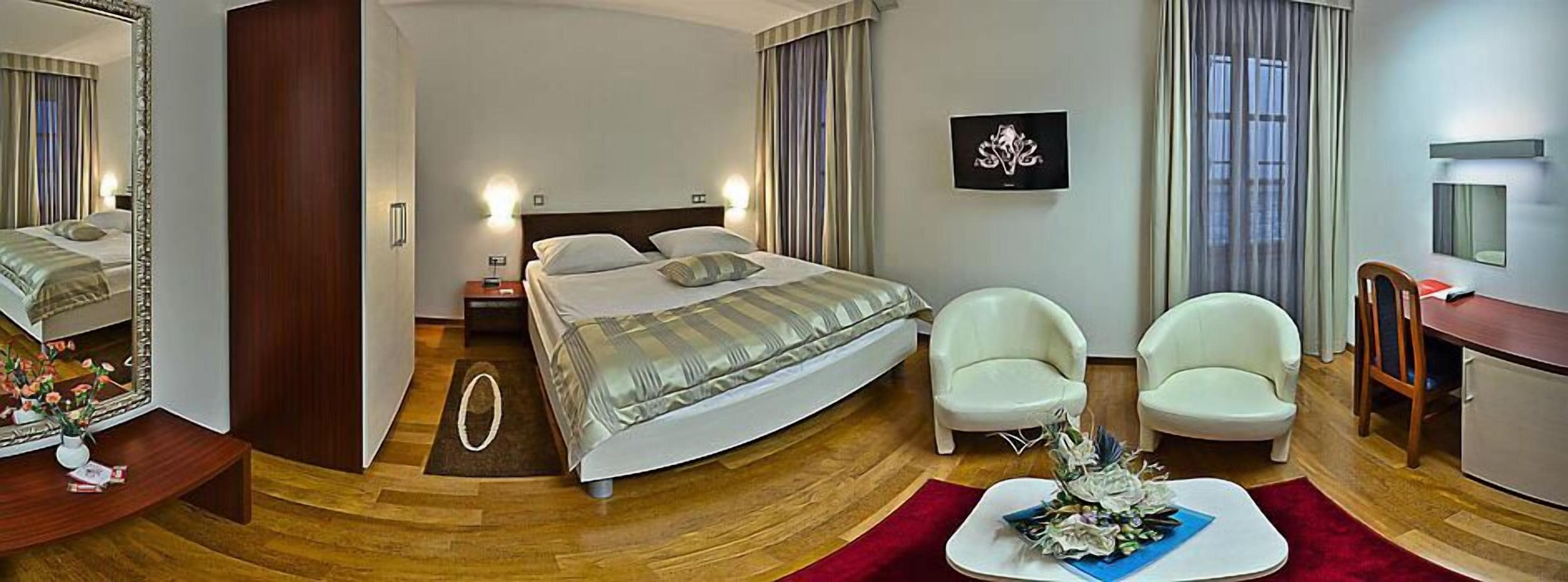 A hotel room with a bed, two white armchairs, a desk, a mirror, a TV, and a red rug.