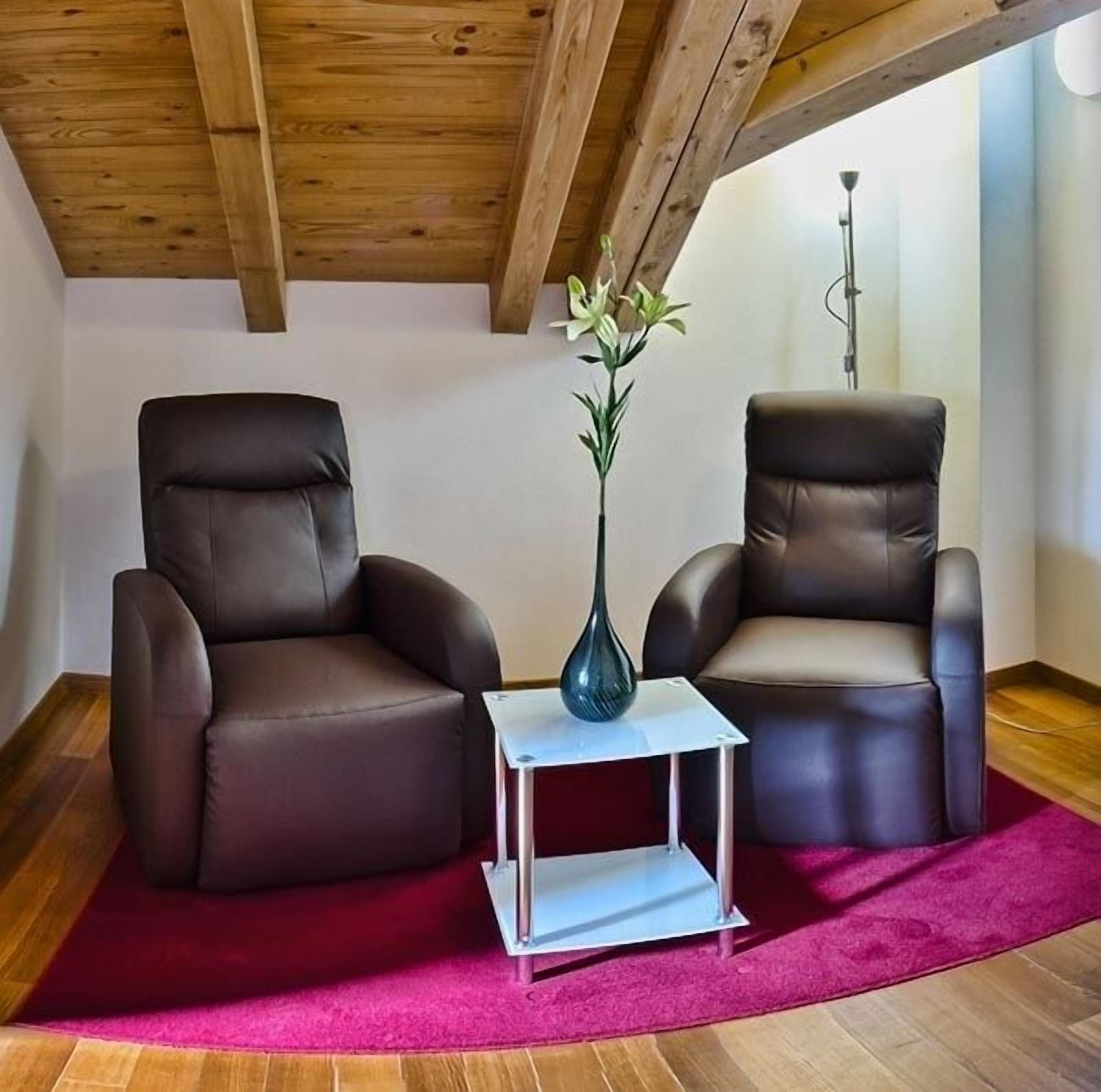 Dos sillones de cuero, una mesita blanca y un florero con flores en una habitación con vigas de madera y alfombra roja.