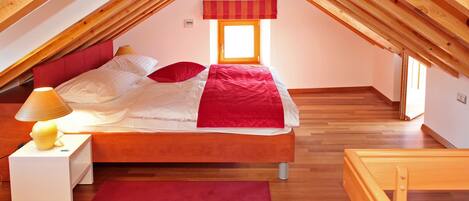 Un dormitorio con techo inclinado, una cama con ropa de cama roja, una lámpara de noche y una ventana con un valance a rayas.