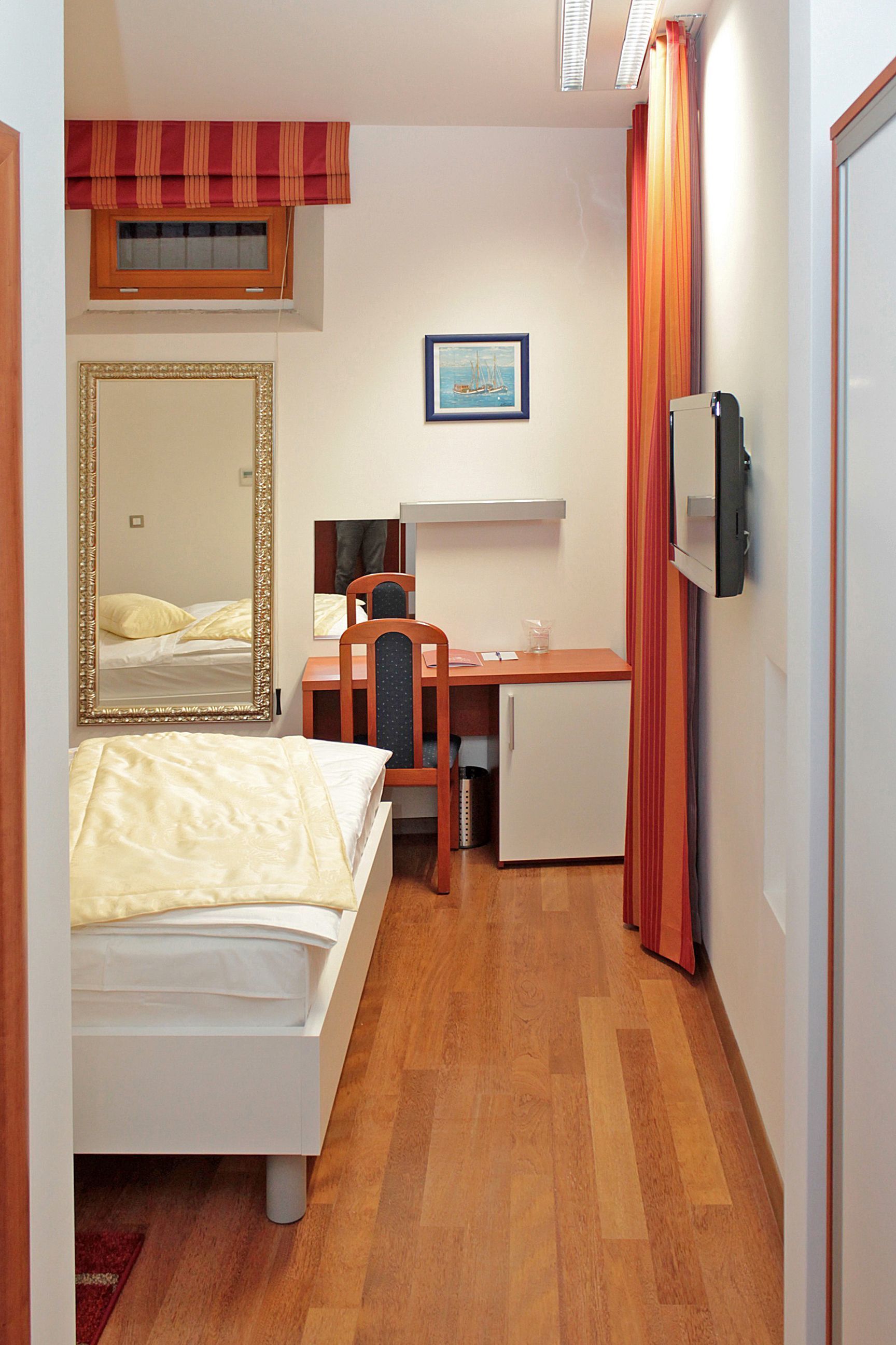A compact hotel room with a bed, a desk with a chair, a small refrigerator, a mirror, and a window with curtains.