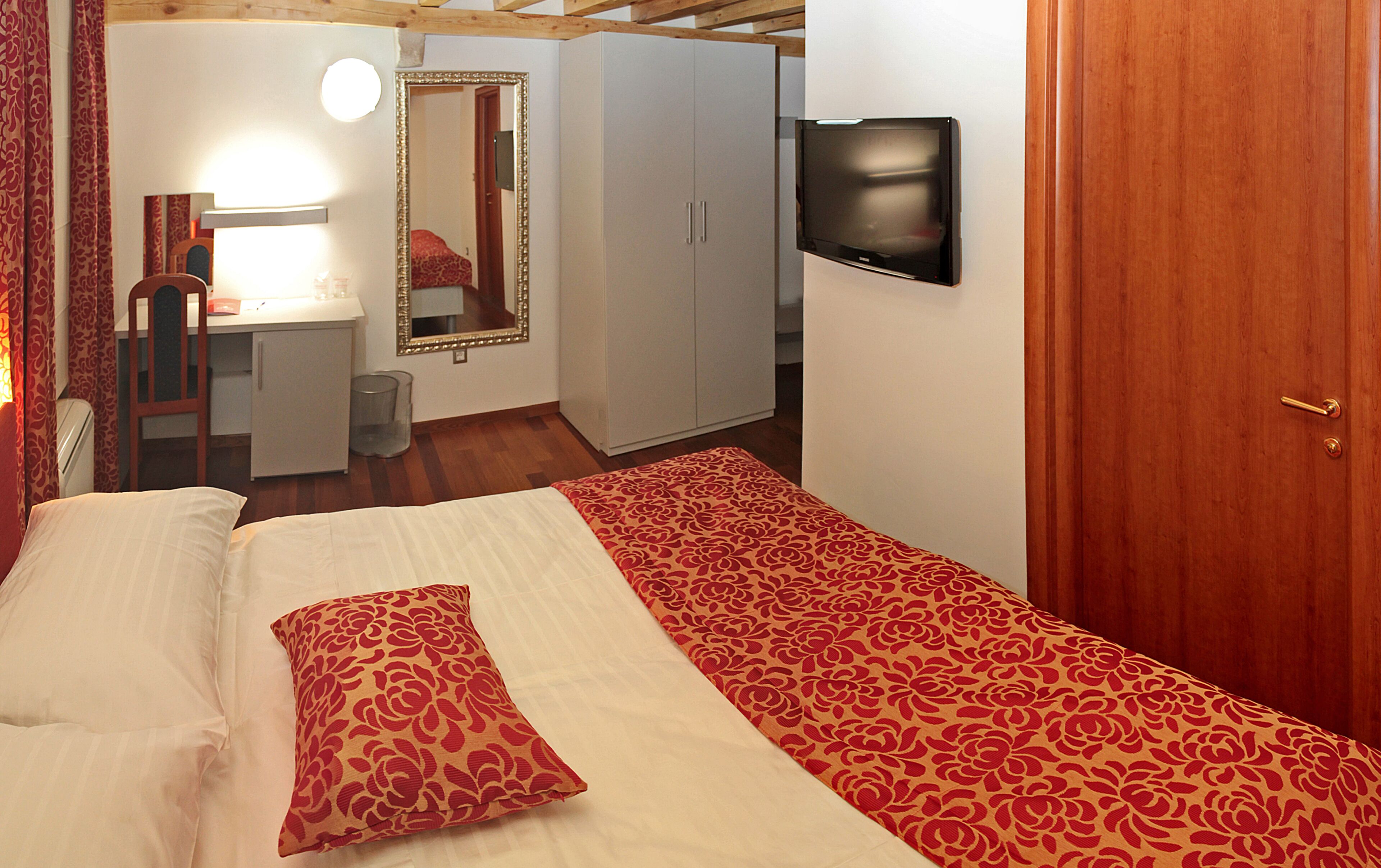 A bedroom with a bed, a desk, a chair, a mirror, and a television.