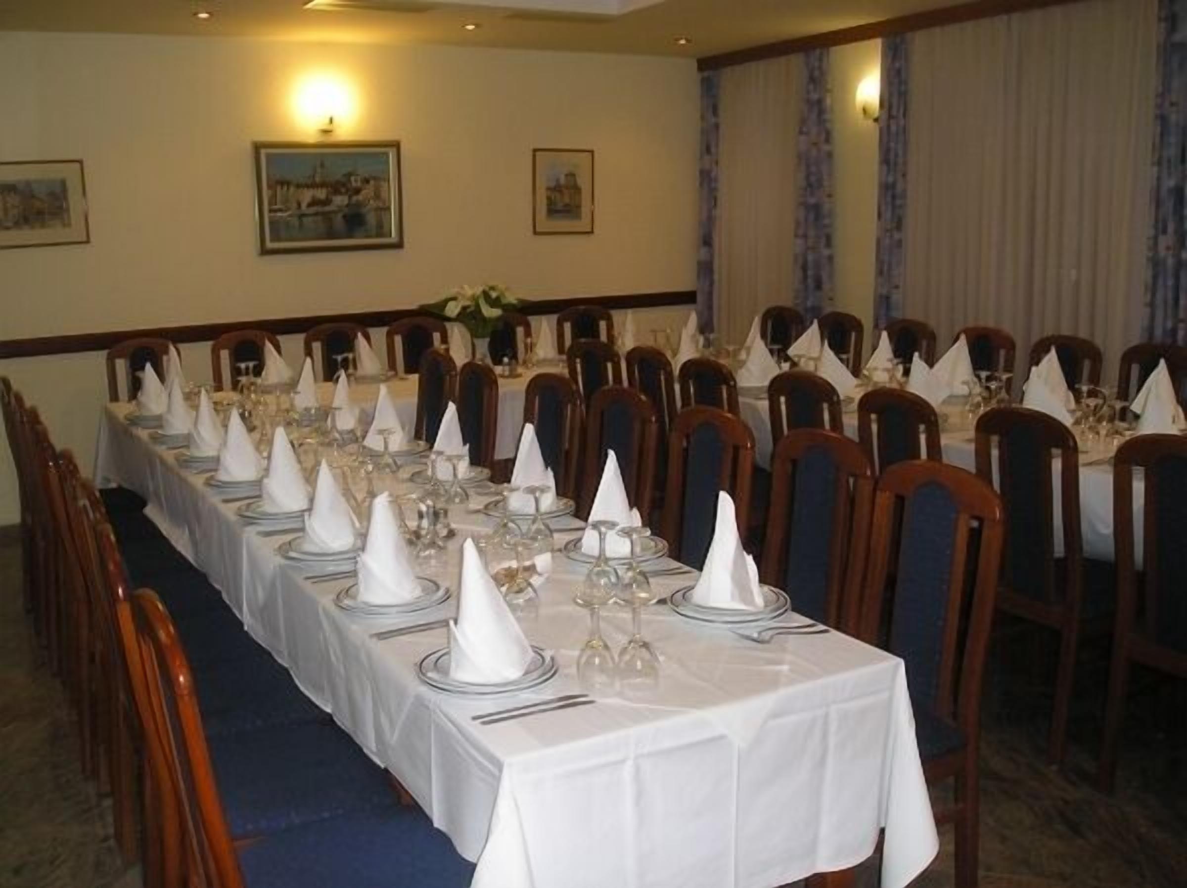 Una larga mesa de comedor cubierta con manteles blancos, copas y servilletas dobladas, rodeada de sillas de madera.